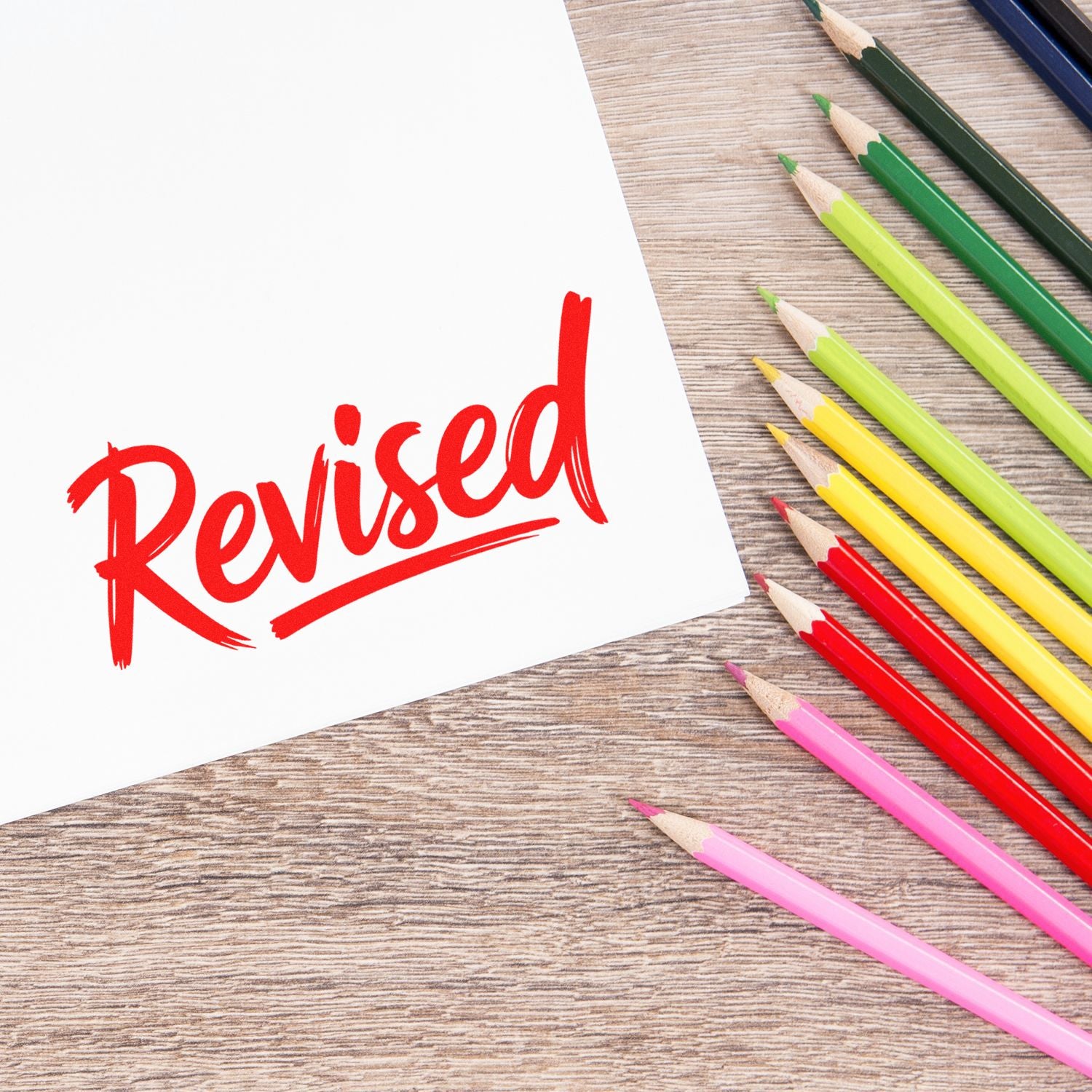 Red Revised rubber stamp on white paper with colored pencils, signaling document review and approval.