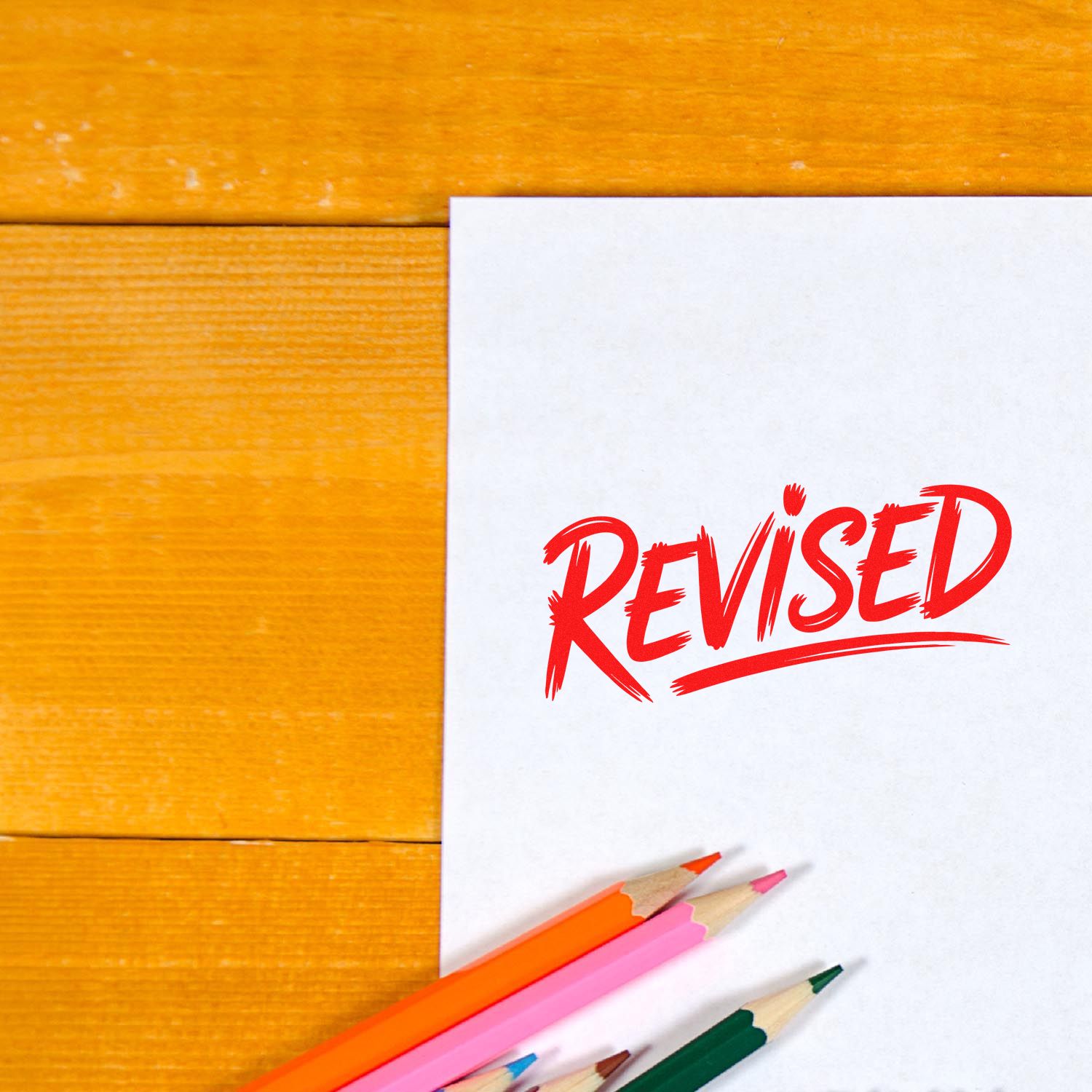 Revised rubber stamp on document with colored pencils, marking edits on a wooden desk.