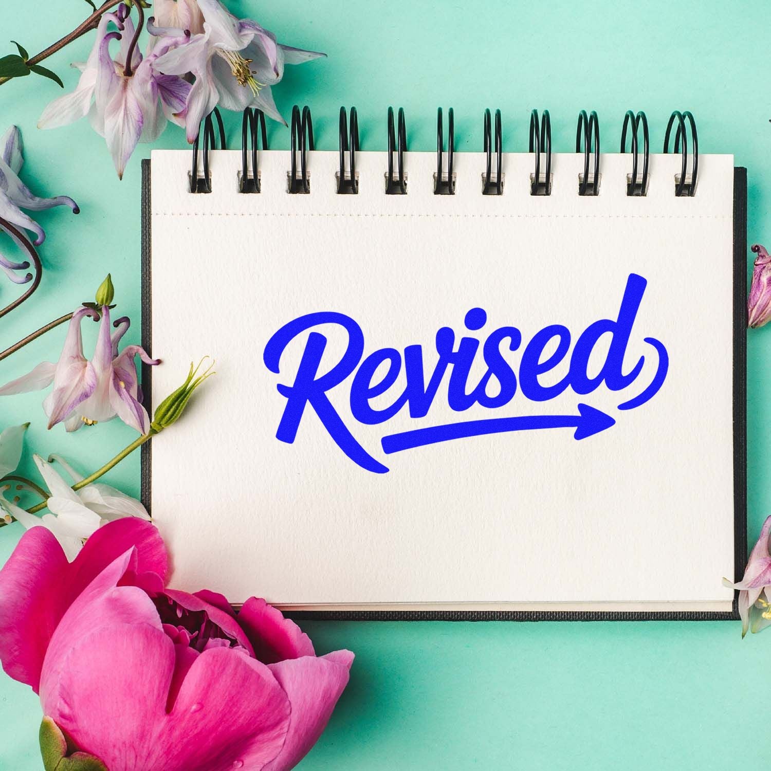 Blue Revised rubber stamp on a spiral notebook with floral accents, ideal for document approval and editing.