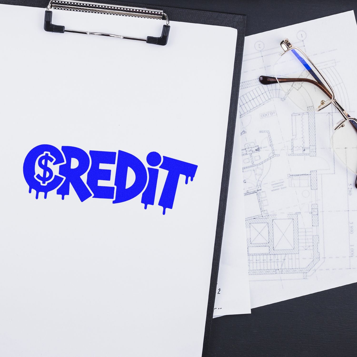 Custom rubber stamp proof with blue CREDIT stamp on clipboard beside eyeglasses and blueprint drawings.