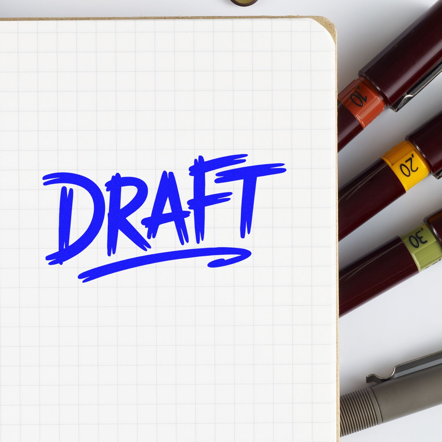 Blue DRAFT rubber stamp impression on notebook page beside colorful markers, for document review and office labeling.