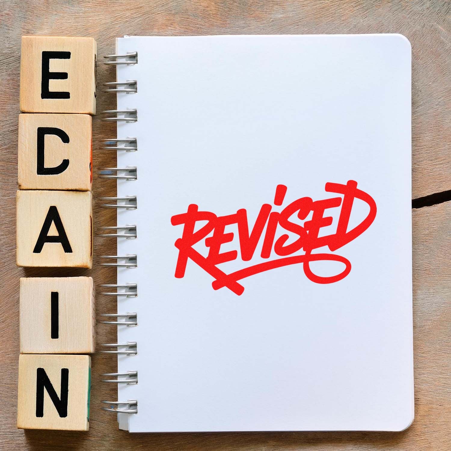 Red REVISED rubber stamp on notebook beside wooden alphabet blocks, showing edited or approved document status.