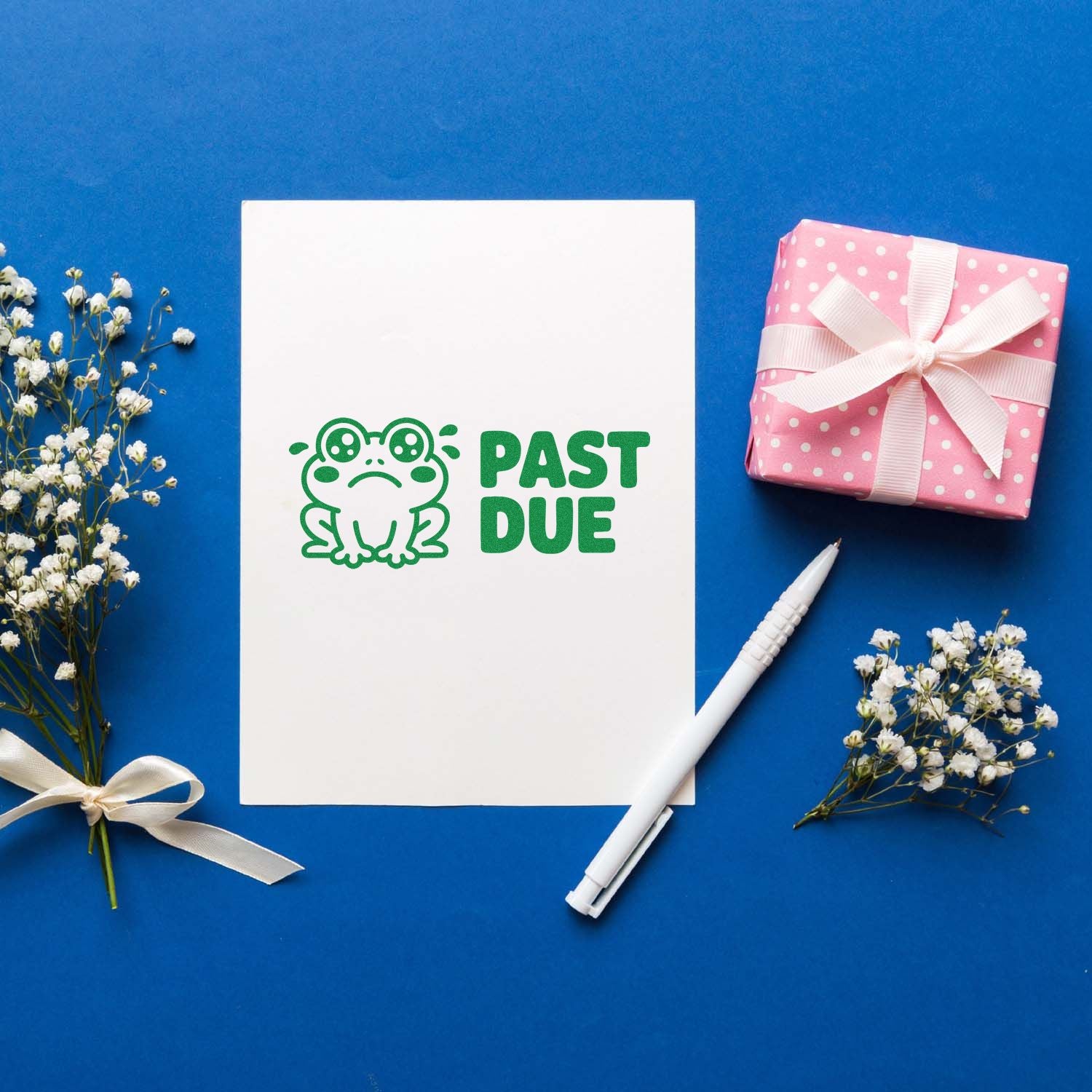 Past Due frog rubber stamp design on a white card, styled with a pen, gift box, and flowers on a blue background.