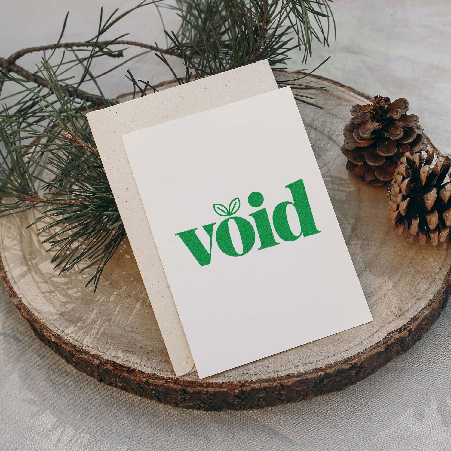 Custom rubber stamp design on a holiday card with green void lettering, pine branches, and pinecones on a rustic wood slice.