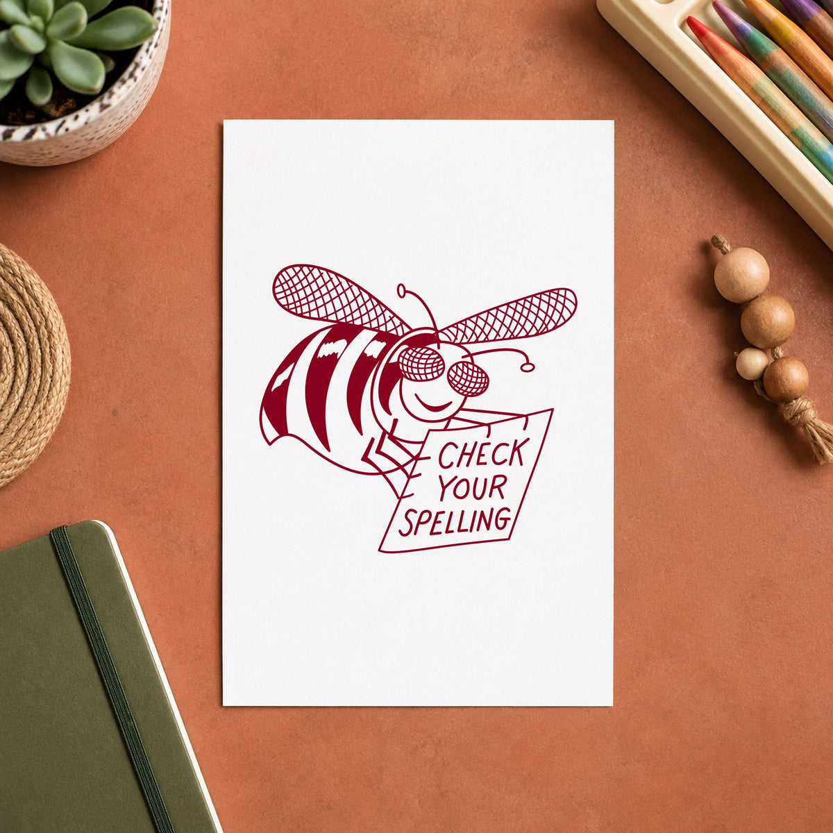 Check Your Spelling Bee Rubber Stamp impression in burgundy ink on white paper in a styled craft setting.