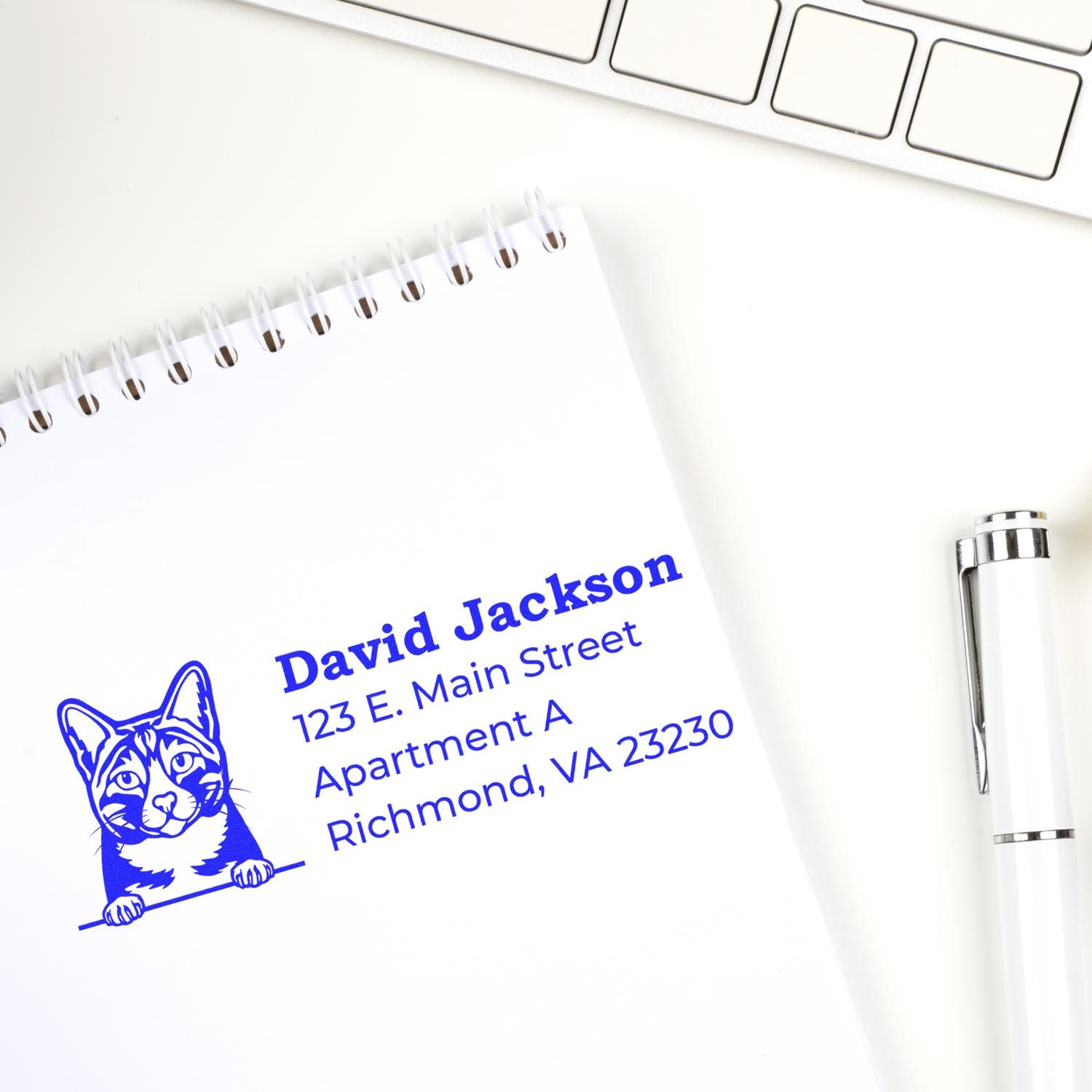 Custom rubber stamp with cat design, blue ink return address imprint on notebook beside pen and keyboard.