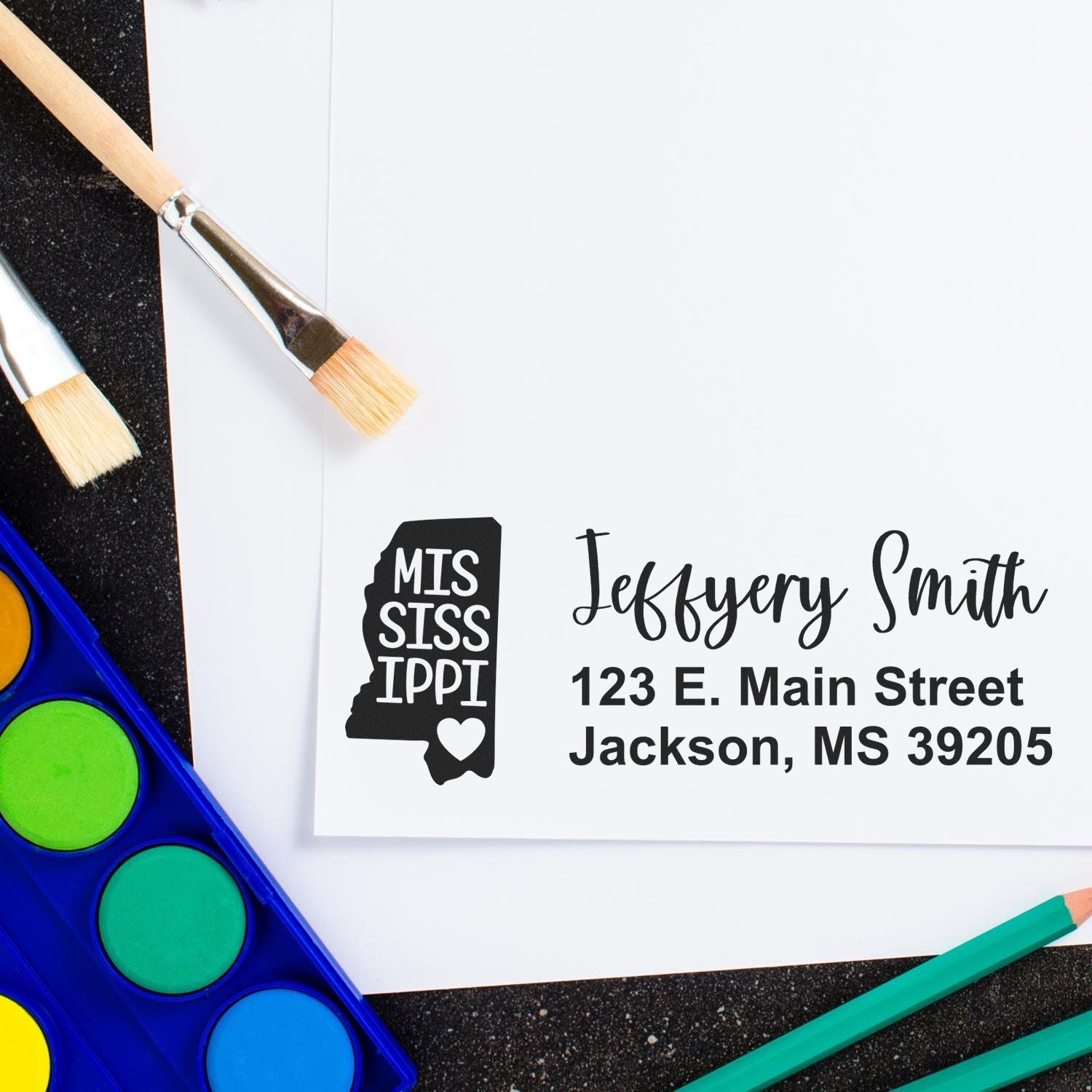 Custom Mississippi rubber stamp with personalized return address for mail, shown with art supplies.