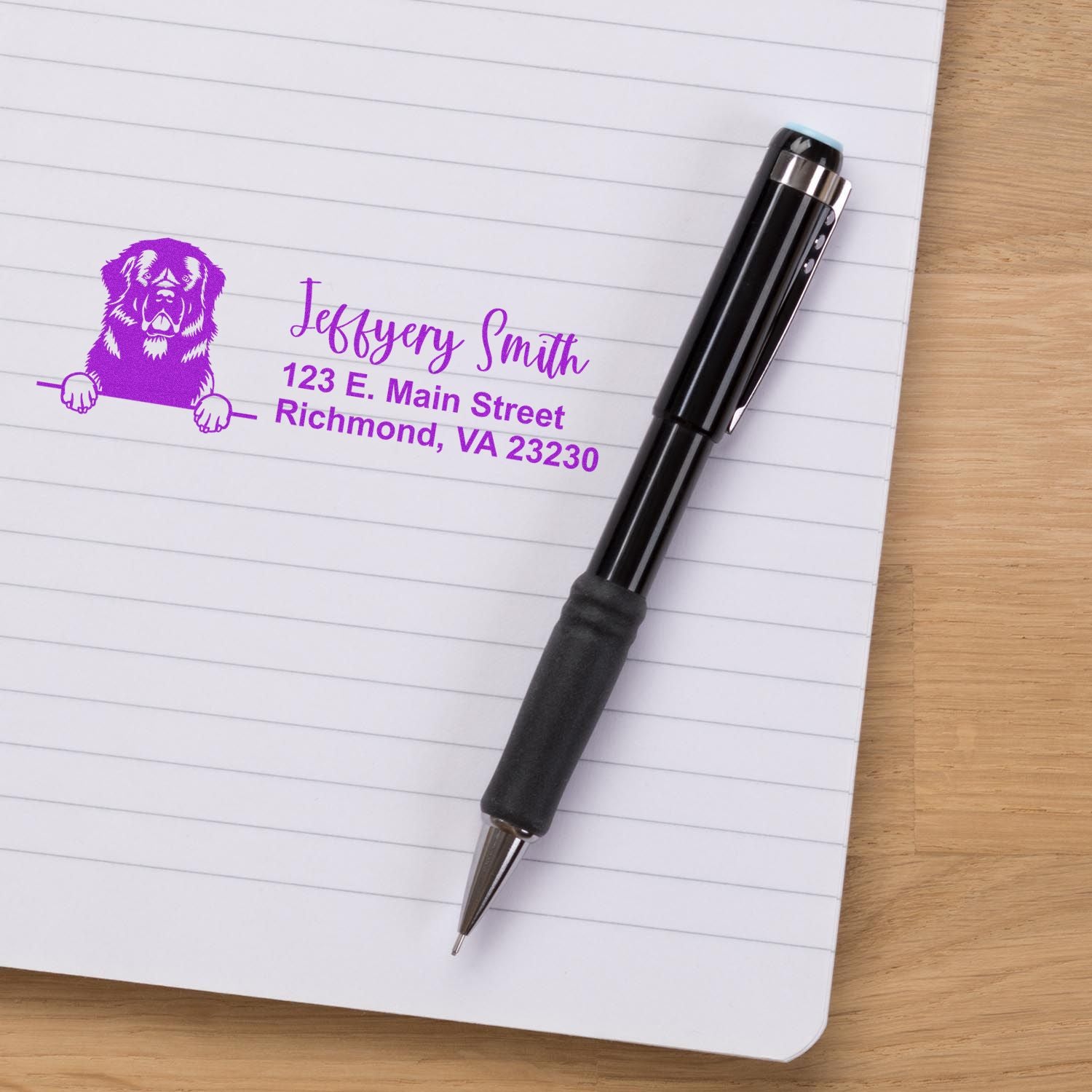 Personalized rubber address stamp with dog logo and purple return address imprint on lined notebook beside a pen.