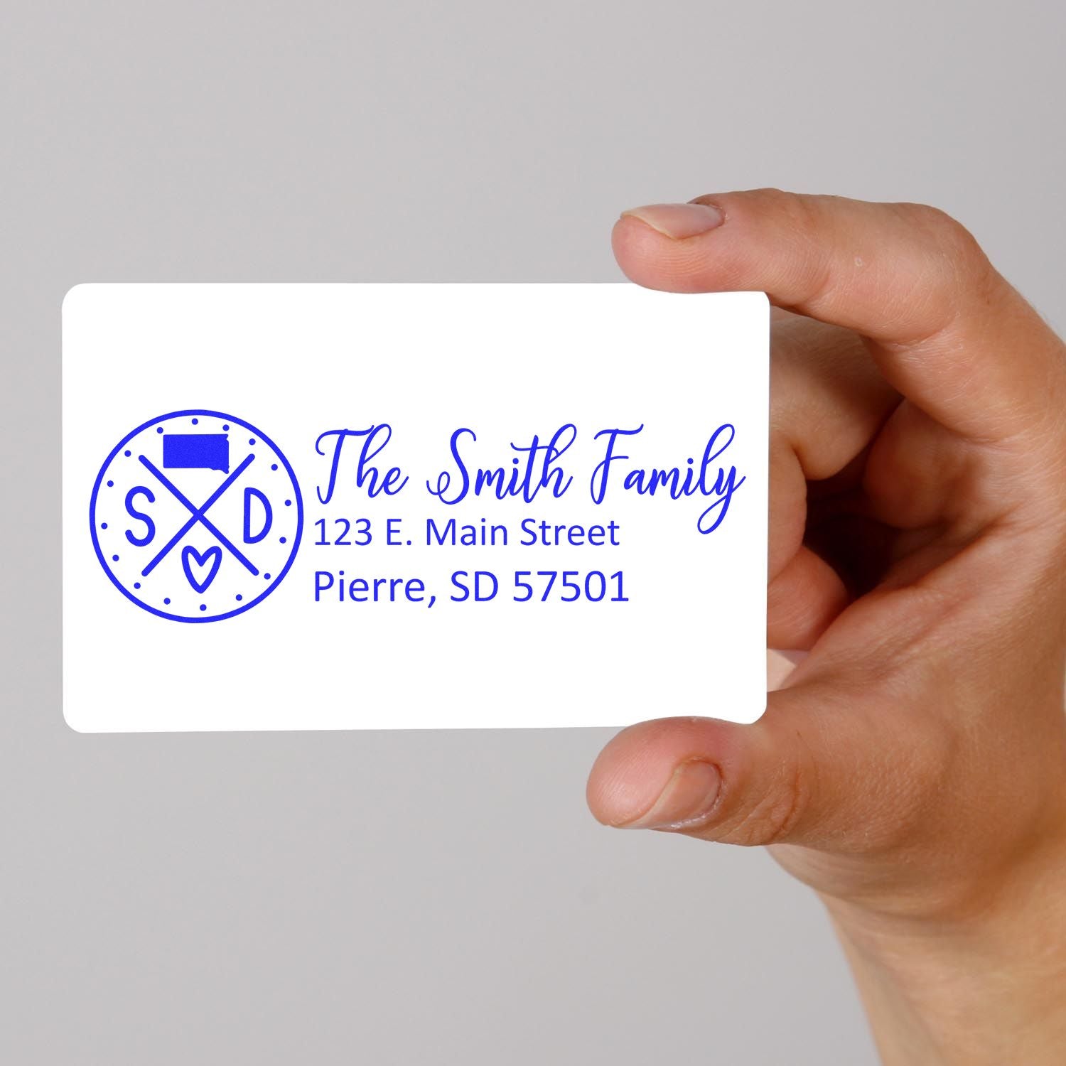 Hand holding custom family return address rubber stamp design with monogram, name, and address in blue ink.