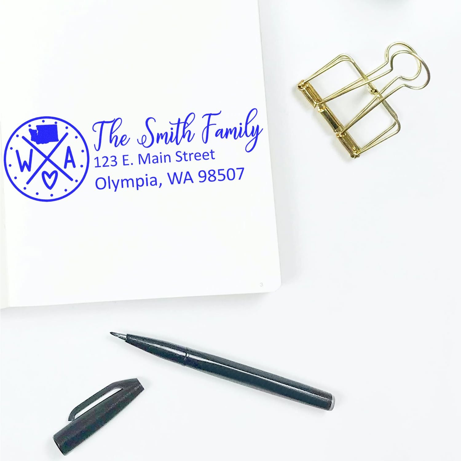 Personalized return address rubber stamp with family name and address imprint, shown with pen and clip.