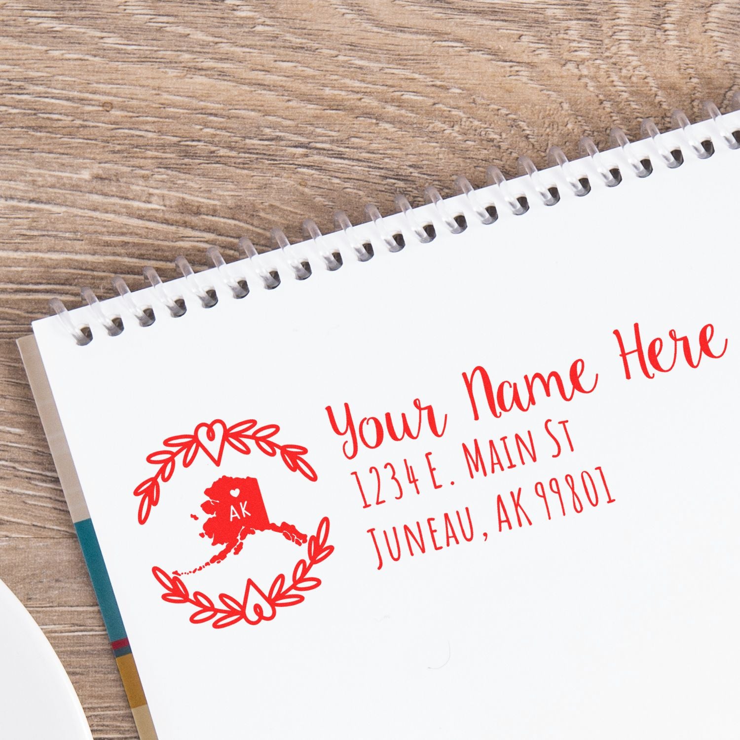 Personalized red rubber return address stamp with Alaska state design, name, and mailing address on notebook paper.