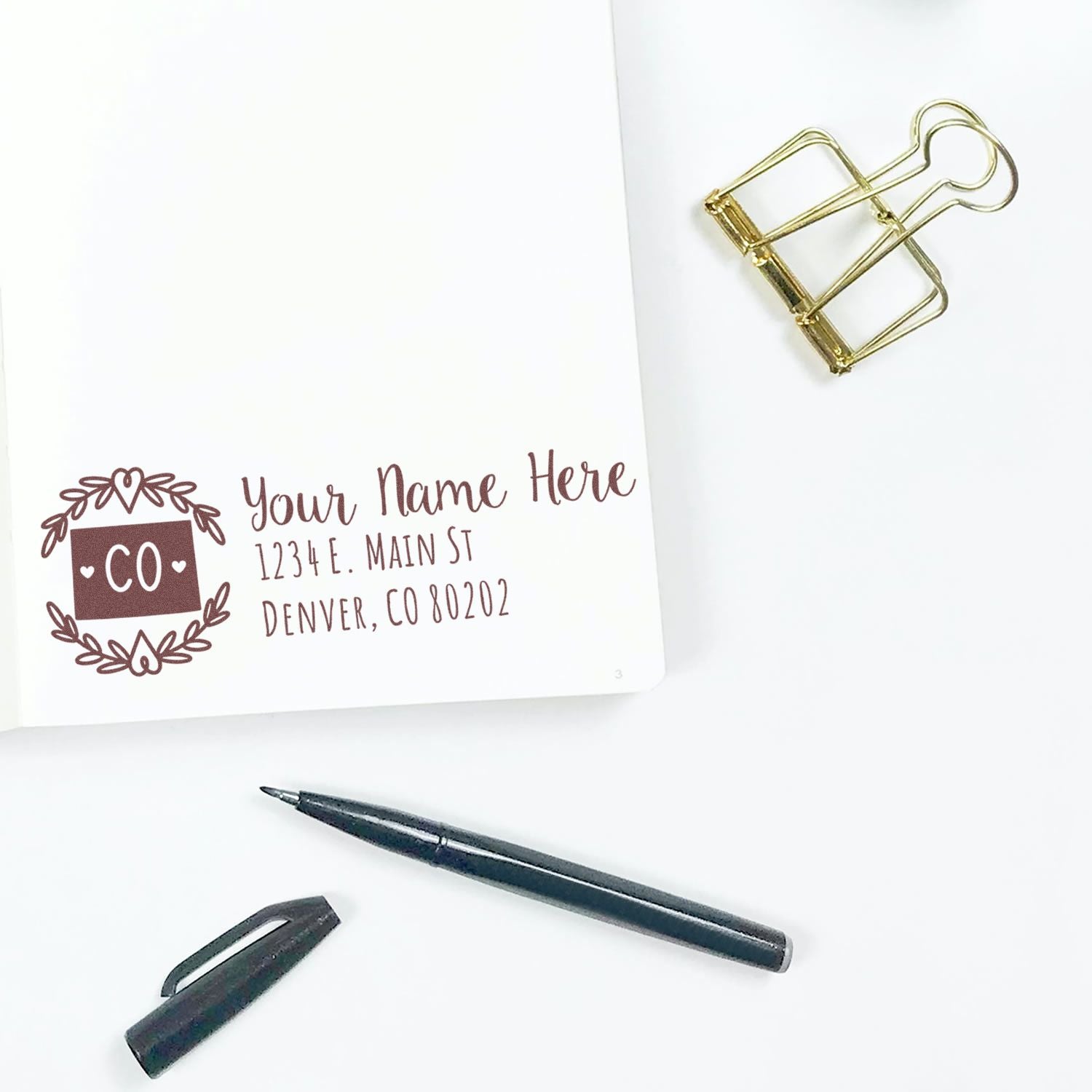 Custom return address rubber stamp design with Colorado address, shown on paper with pen and binder clip.