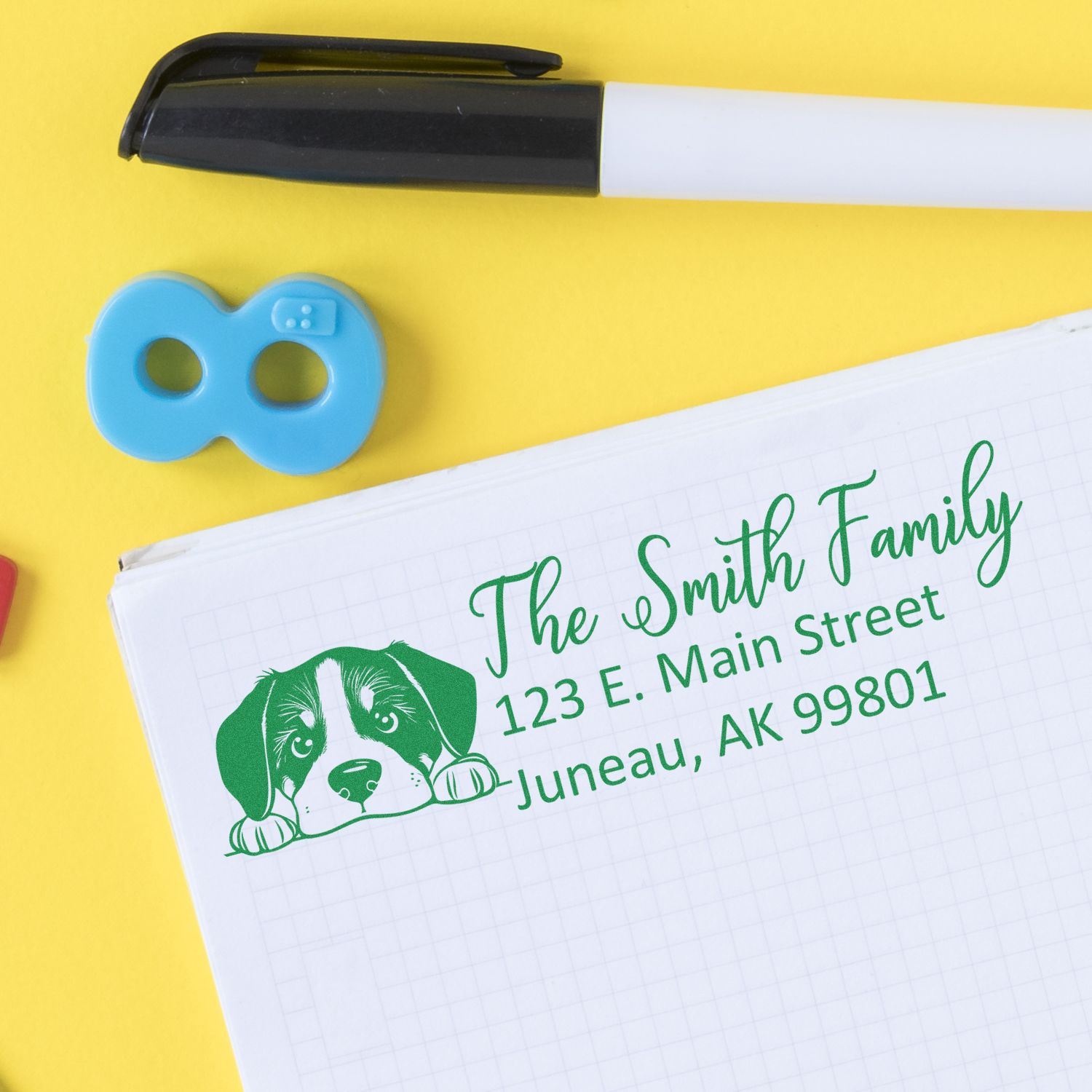 Custom rubber return address stamp with dog illustration, shown stamped on notebook paper beside a marker and stamp case.