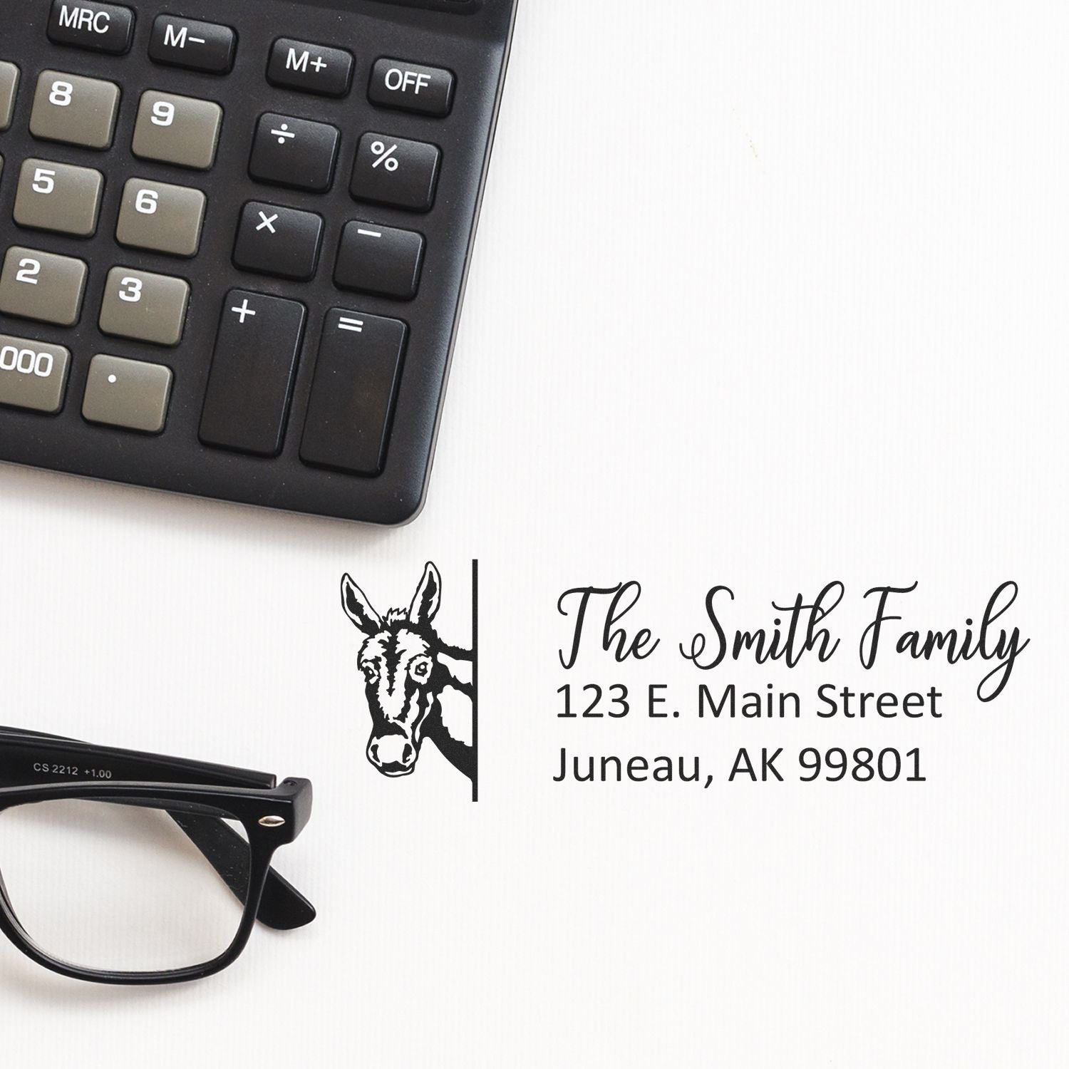 Custom return address rubber stamp with donkey logo, The Smith Family name and Juneau, AK address beside calculator and glasses.