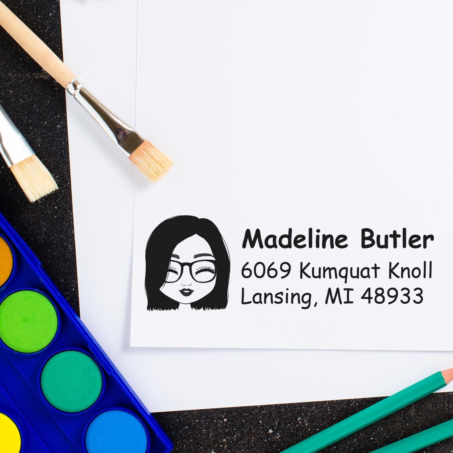Custom return address rubber stamp with illustrated woman logo, shown on paper with art supplies for craft branding.