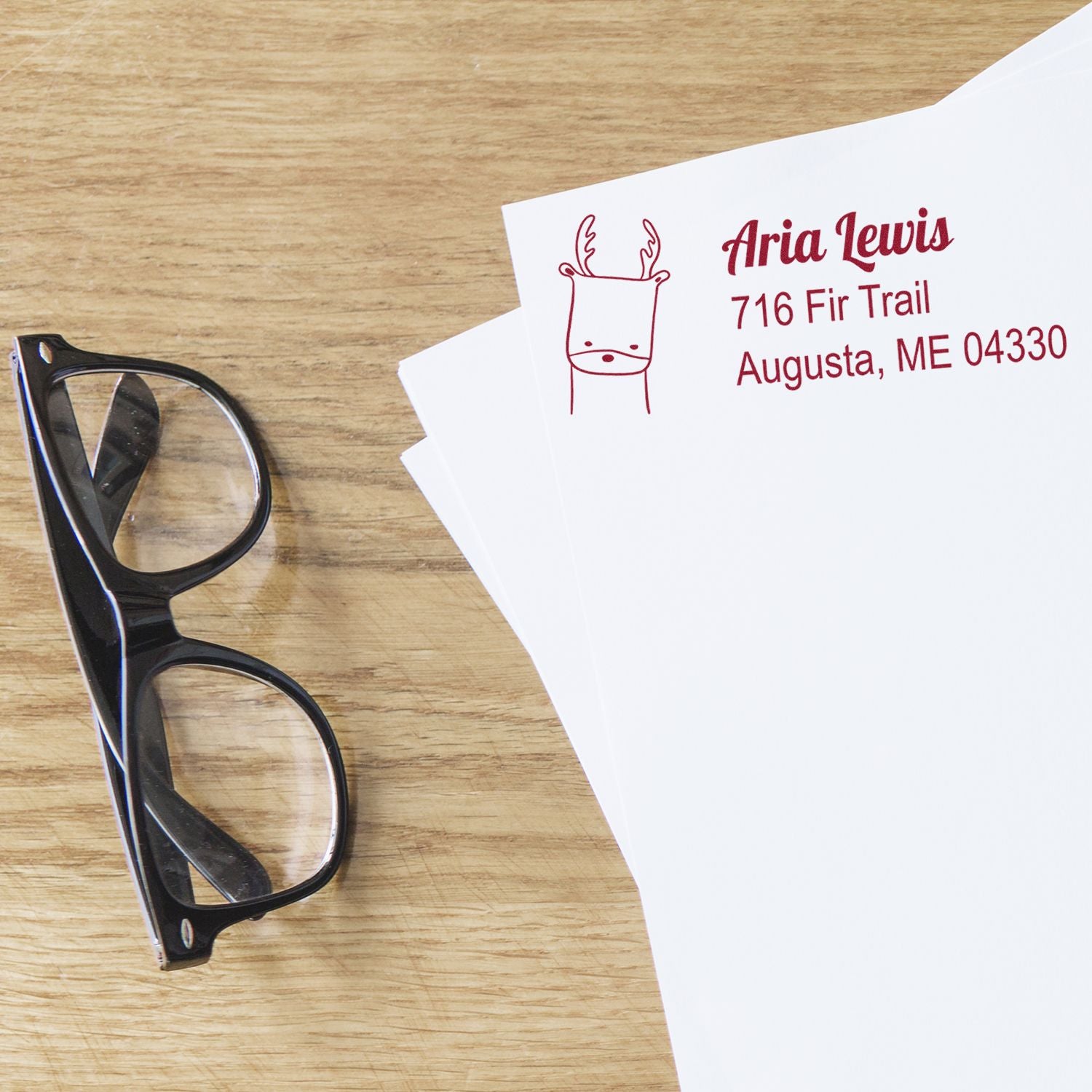 Custom return address rubber stamp with reindeer design on envelopes beside glasses on a wooden desk.