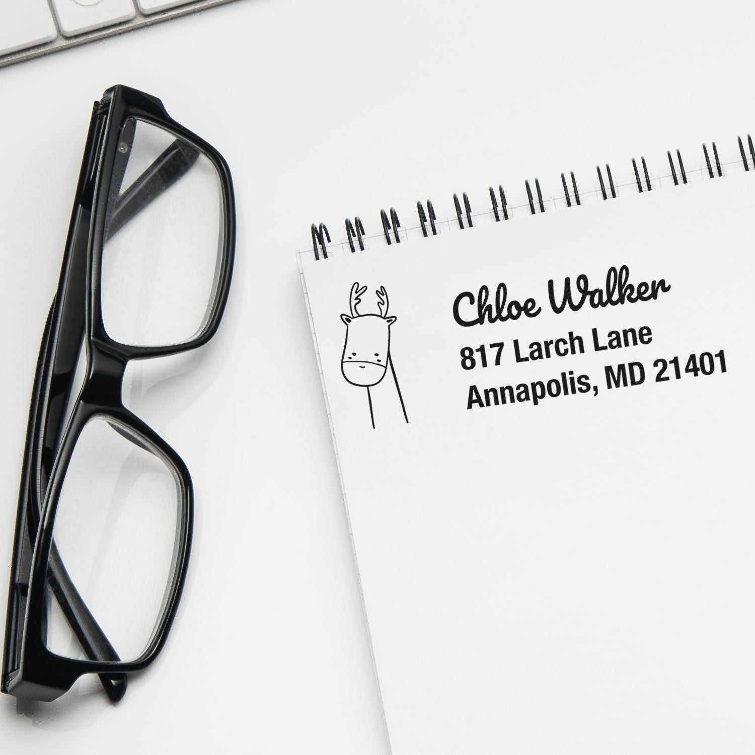 Custom rubber return address stamp with deer icon and personalized name and address on notepad beside eyeglasses.