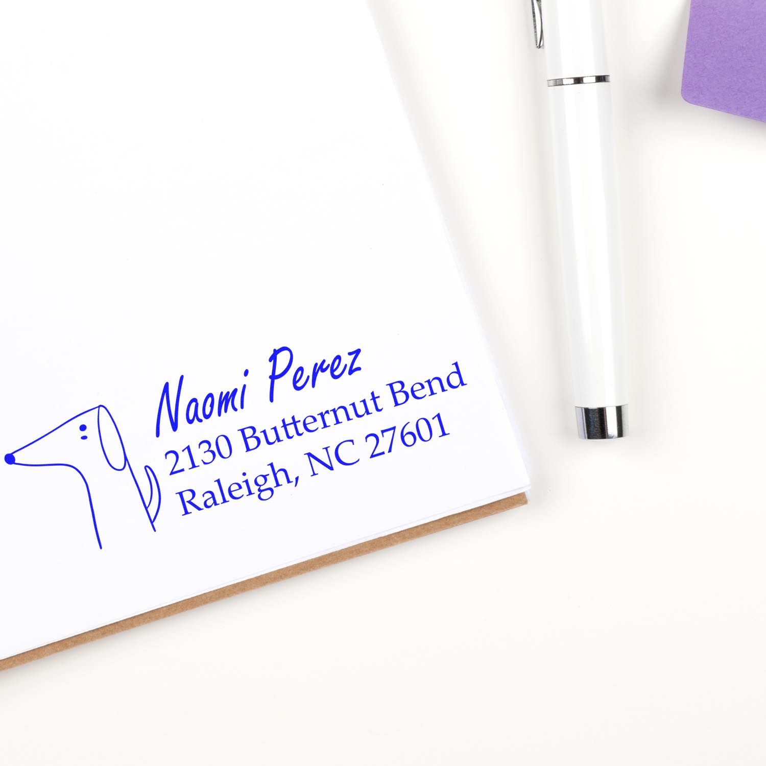 Custom return address rubber stamp impression with name and Raleigh, NC address beside a white pen.
