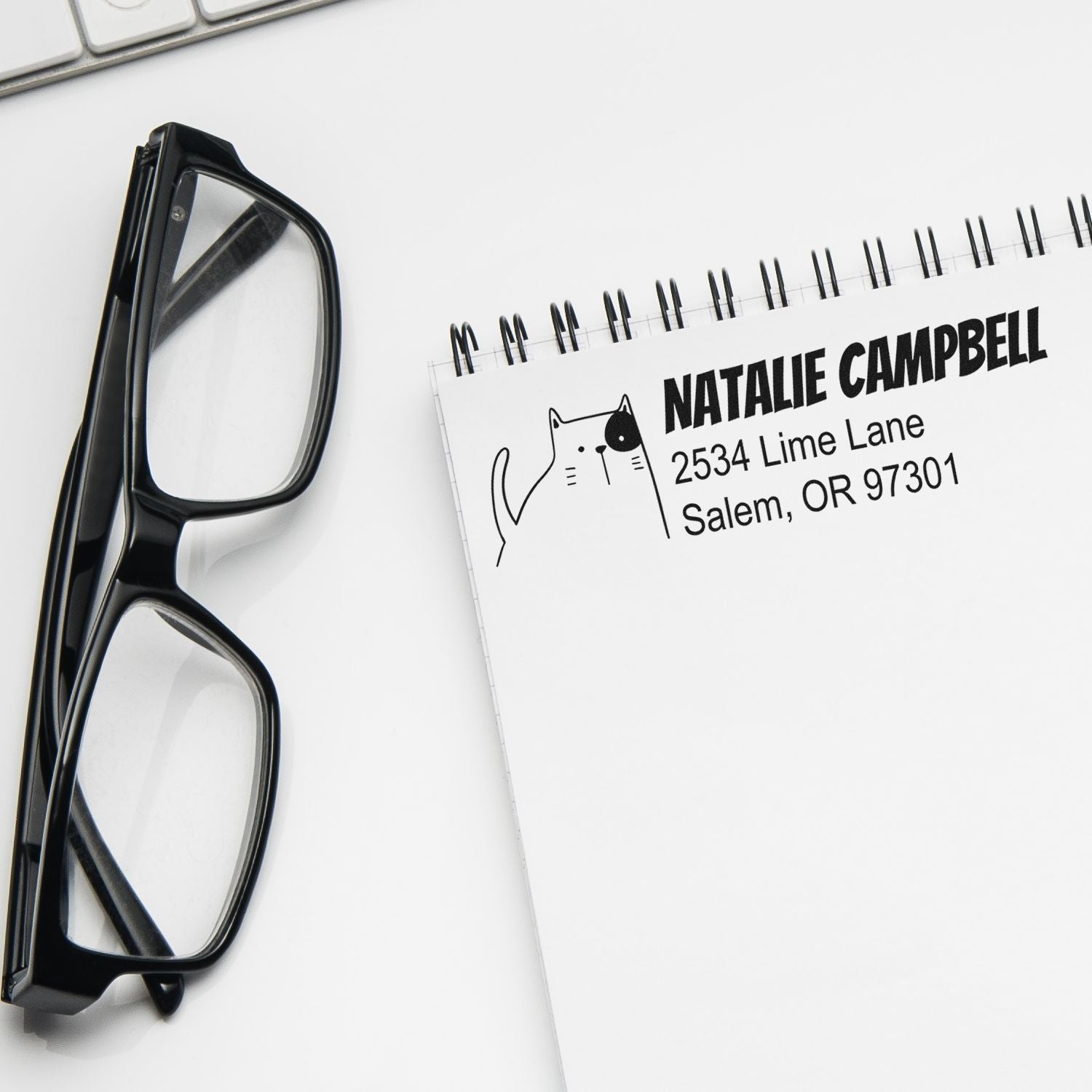Custom address rubber stamp design with cat illustration and name on notebook, beside eyeglasses.