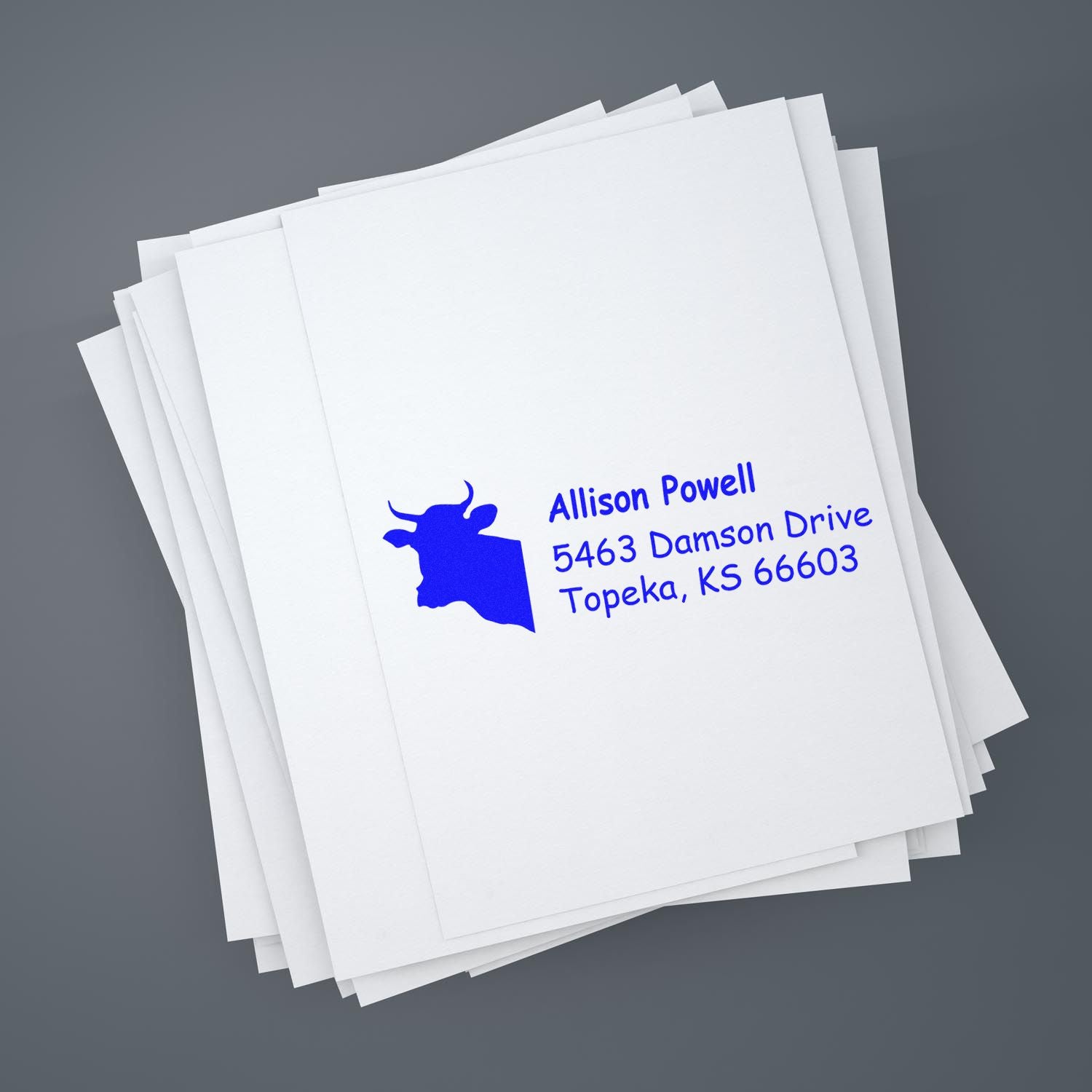 Custom return address rubber stamp with cow logo on stacked white paper, featuring name and Topeka, KS address.