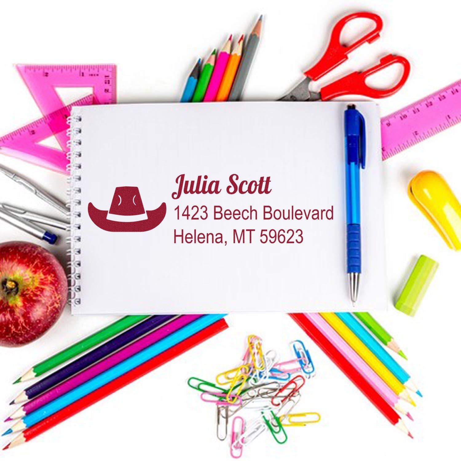 Custom rubber address stamp for personalized mailing, showing name and address on paper with colorful school supplies.