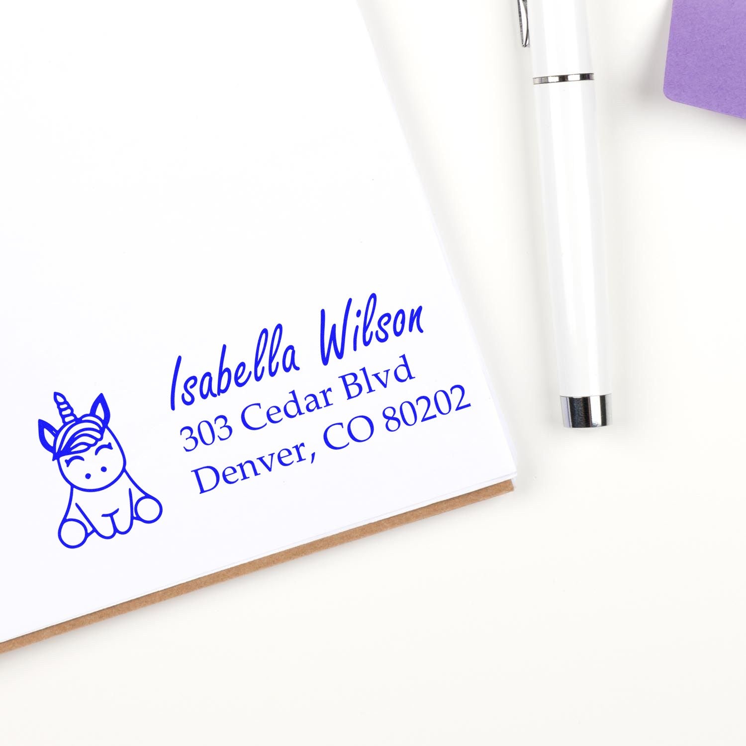 Personalized unicorn address rubber stamp with name and Denver address, shown stamped on a notebook beside a white pen.