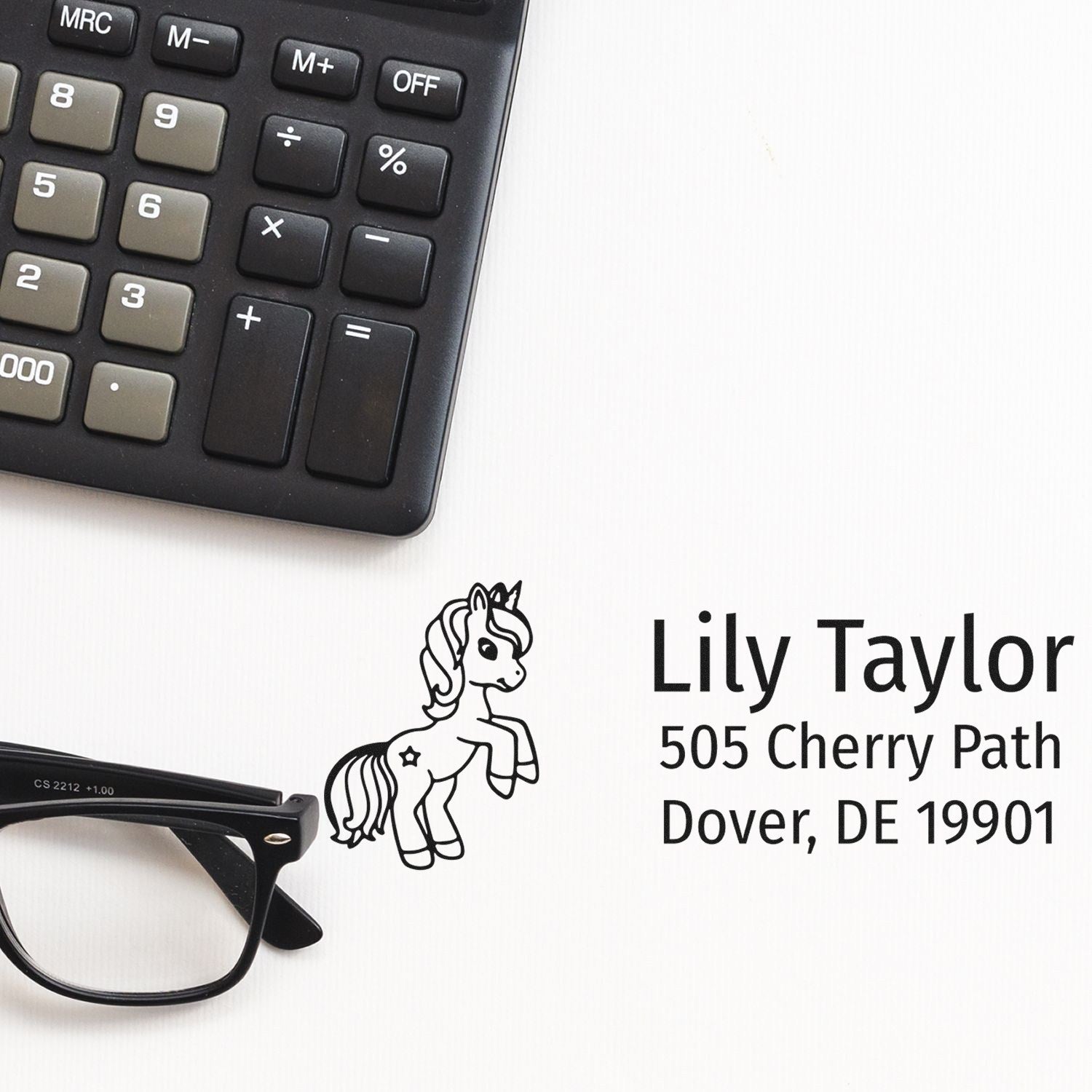 Custom unicorn return address rubber stamp with name and Dover, DE address, shown beside calculator and eyeglasses.
