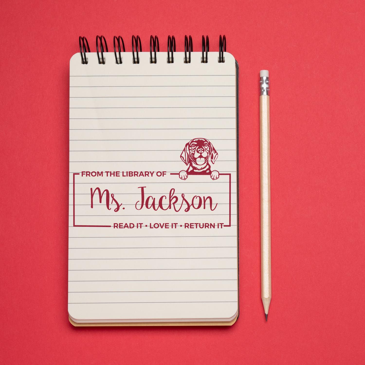 Custom library rubber stamp imprint reading From the Library of Mrs. Jackson with dog design on notebook, red background.