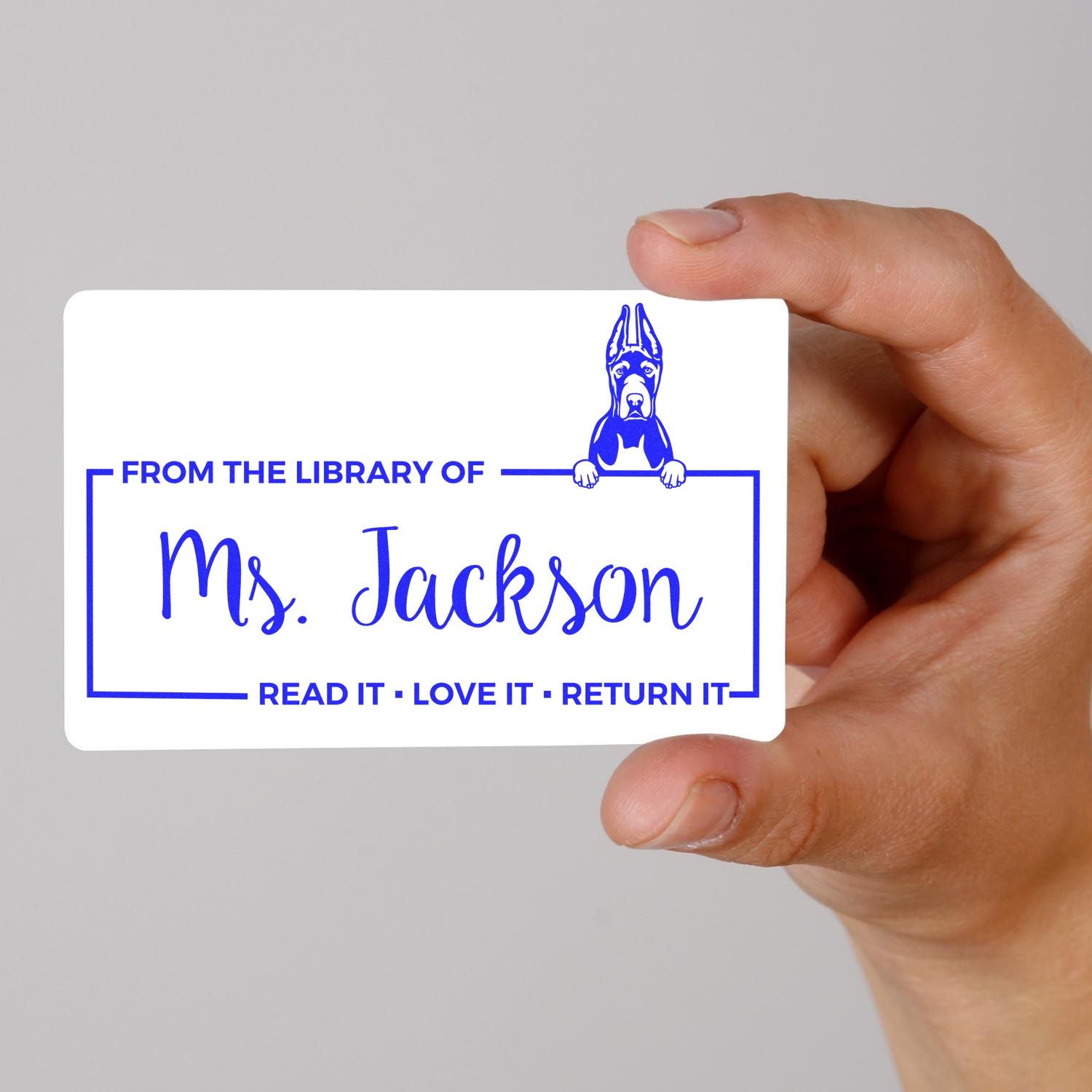 Personalized rubber library stamp with dog design and From the Library of Ms. Jackson book return message, held in hand.