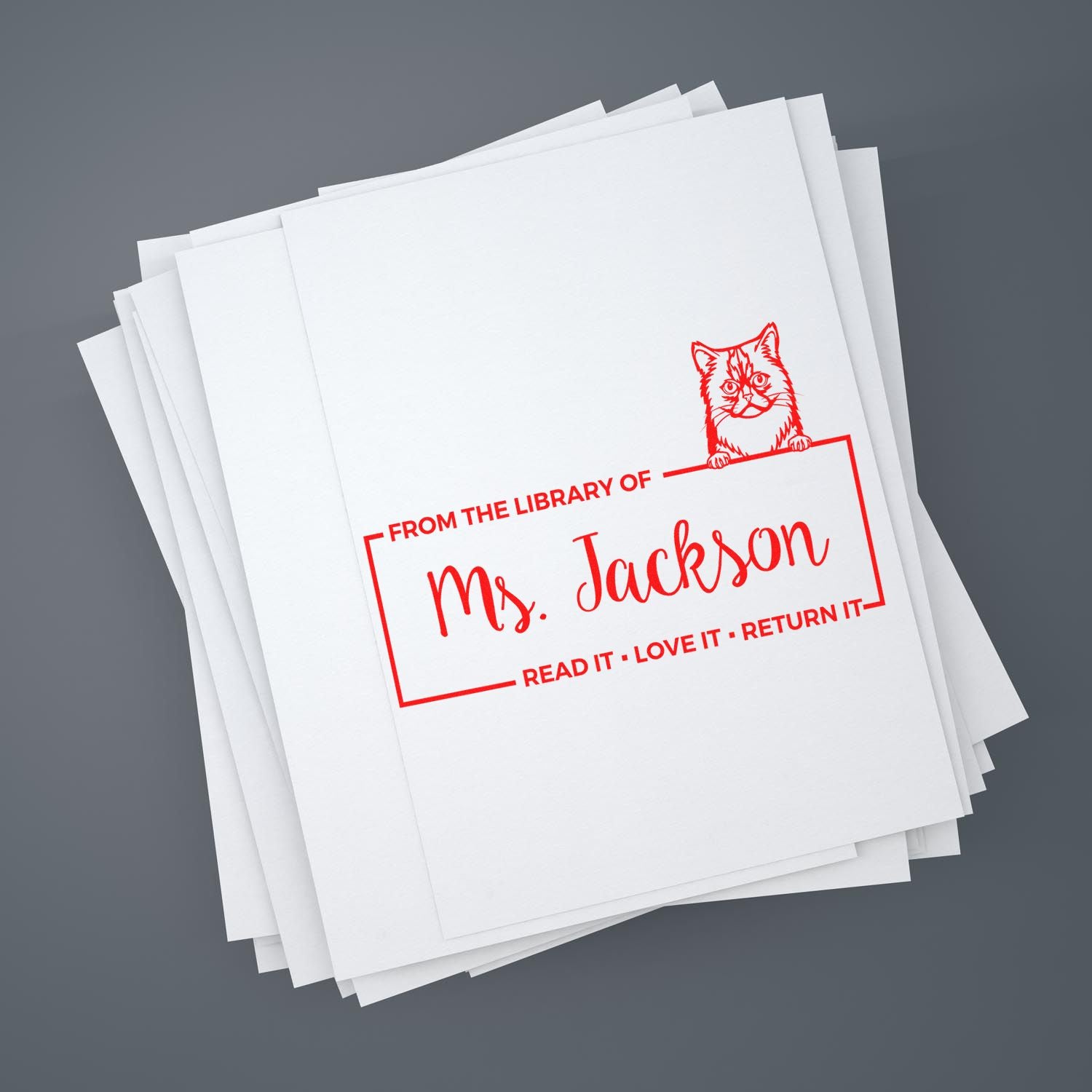 Personalized library rubber stamp for books, featuring cat design and Ms. Jackson name, on stacked papers.
