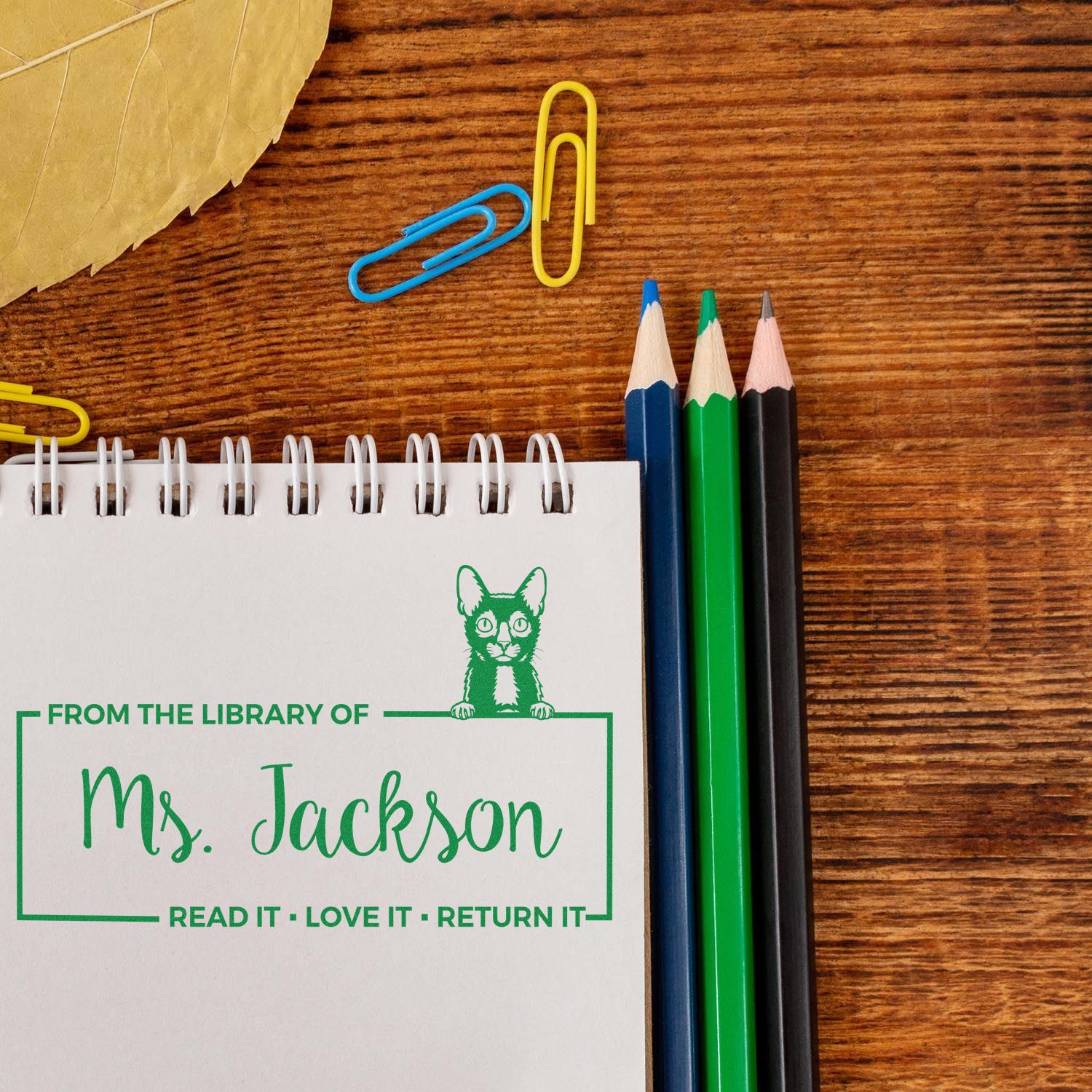 Personalized library rubber stamp on notebook reading From the Library of Ms. Jackson, with pencils and paper clips.