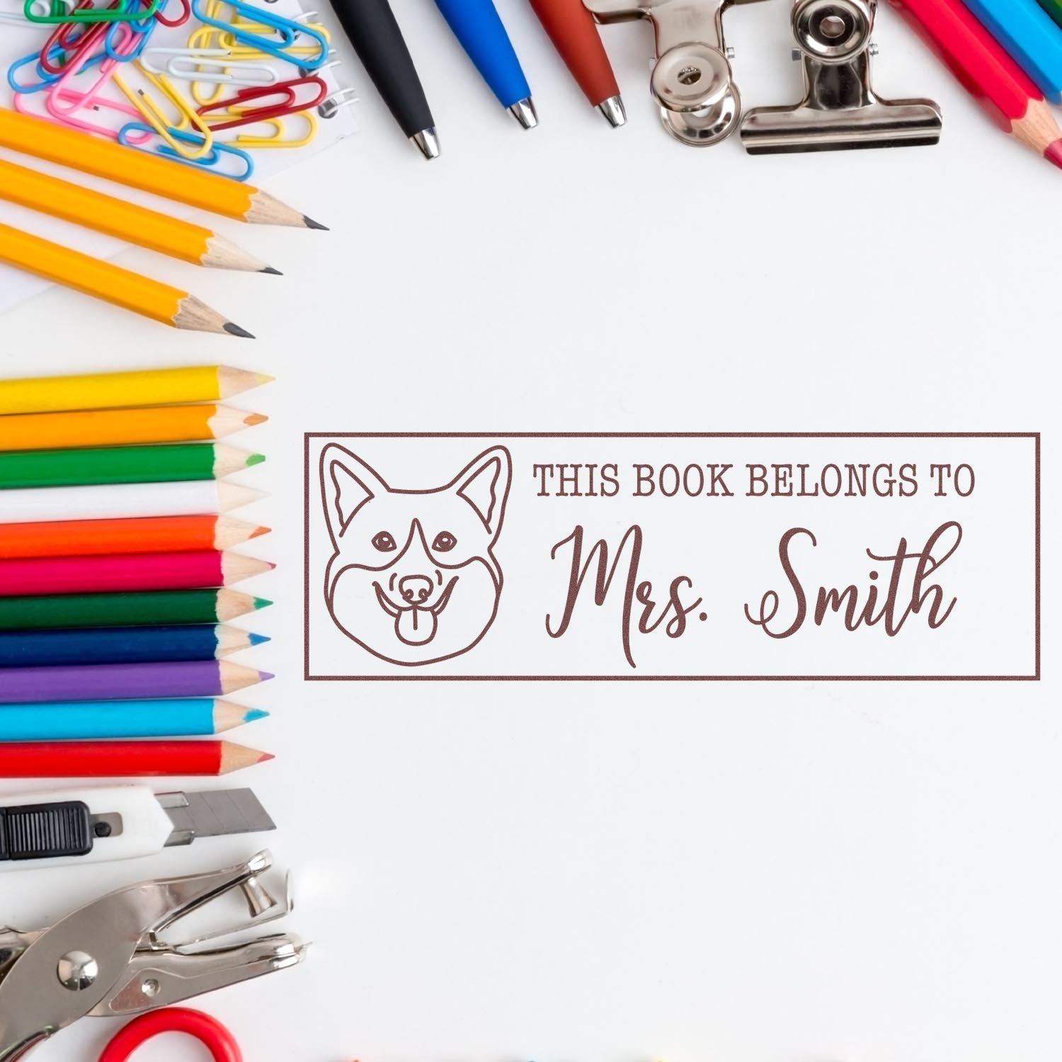 Personalized rubber stamp for books with dog illustration and This Book Belongs to Mrs. Smith, shown with stationery supplies.