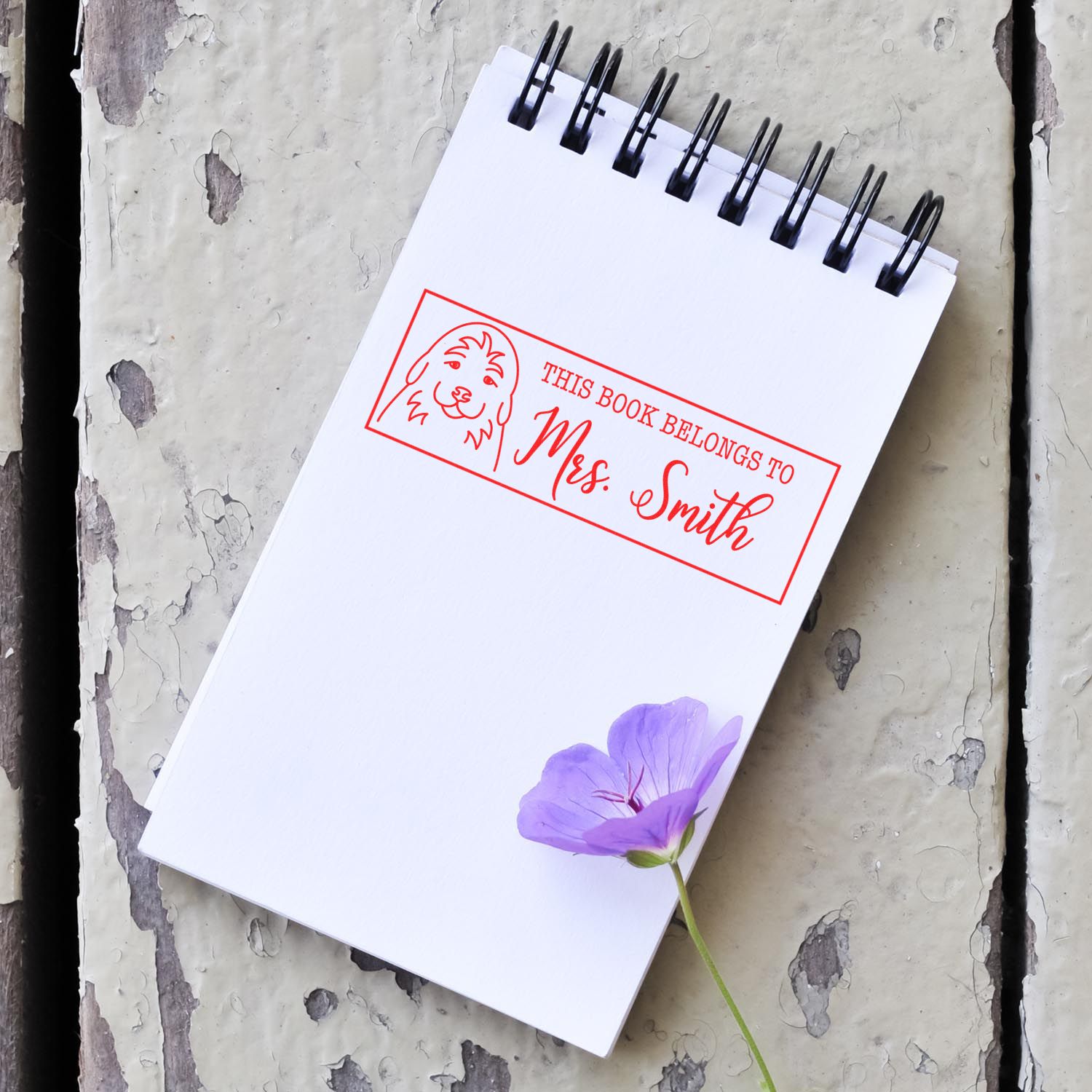 Personalized rubber stamp on notebook reading This Book Belongs to Mrs. Smith with dog icon, ideal for custom library book labels.