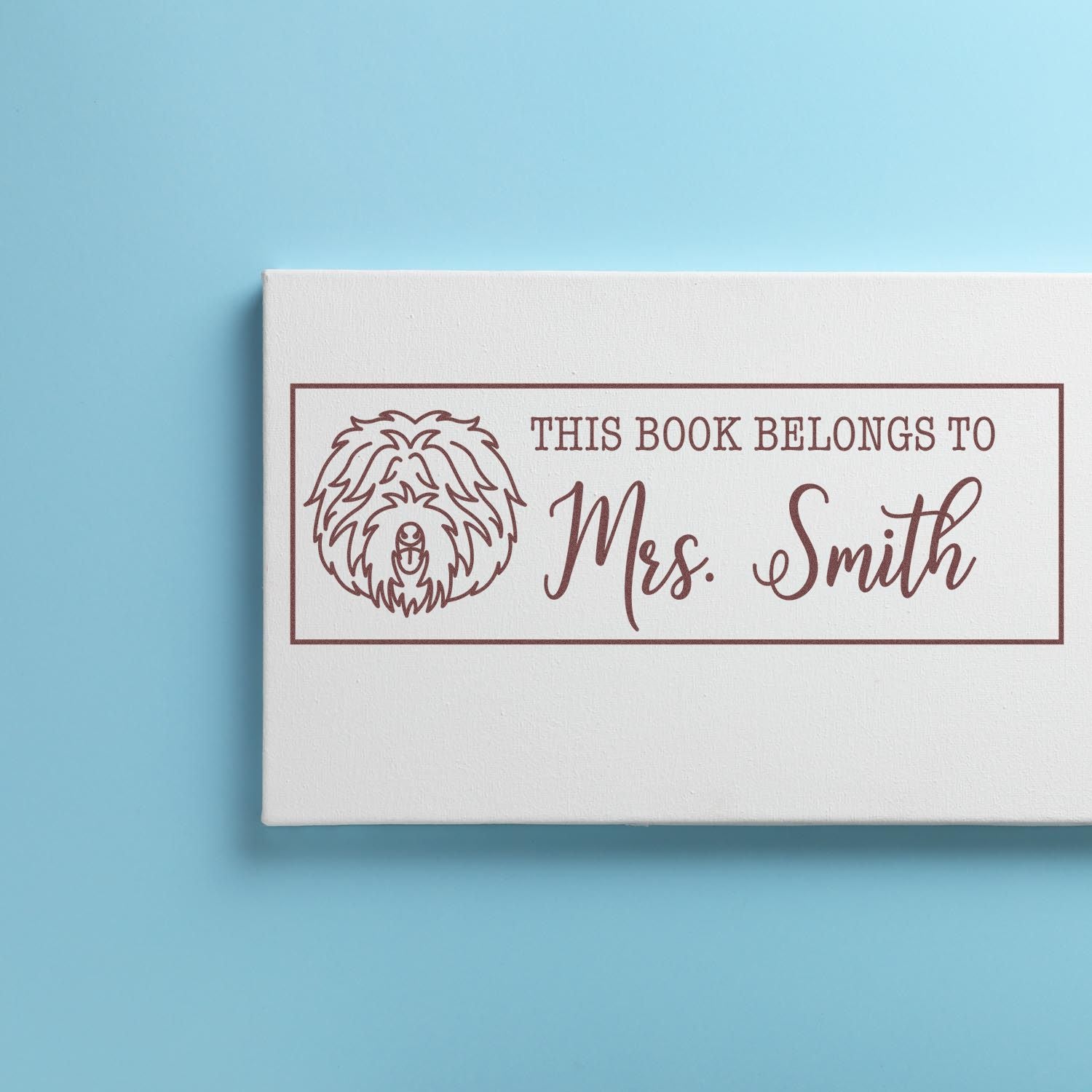Custom rubber stamp on a book cover reading This Book Belongs to Mrs. Smith with a dog icon for personalized library labeling.