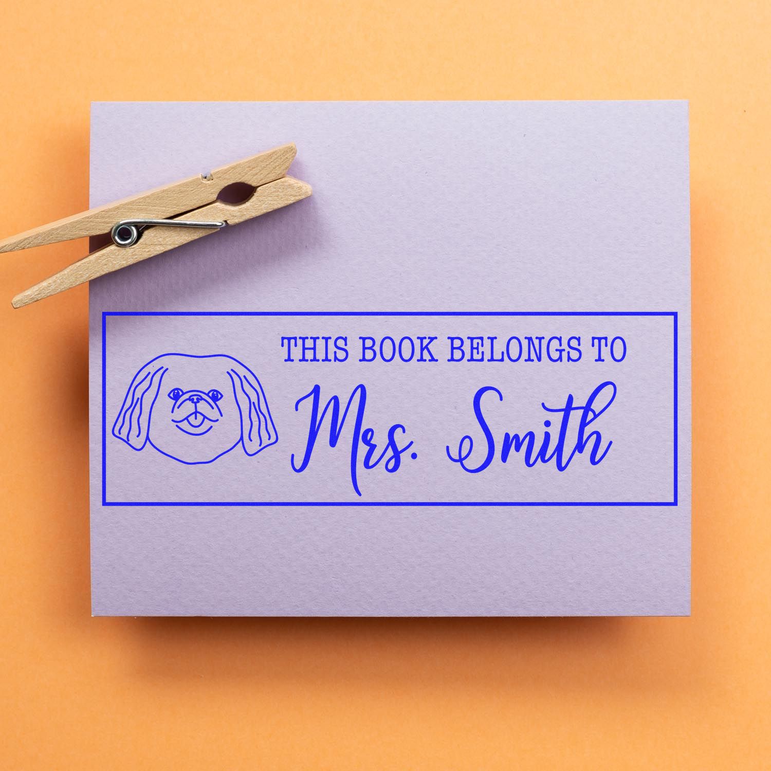 Personalized book rubber stamp with dog illustration and This Book Belongs to Mrs. Smith on purple paper.
