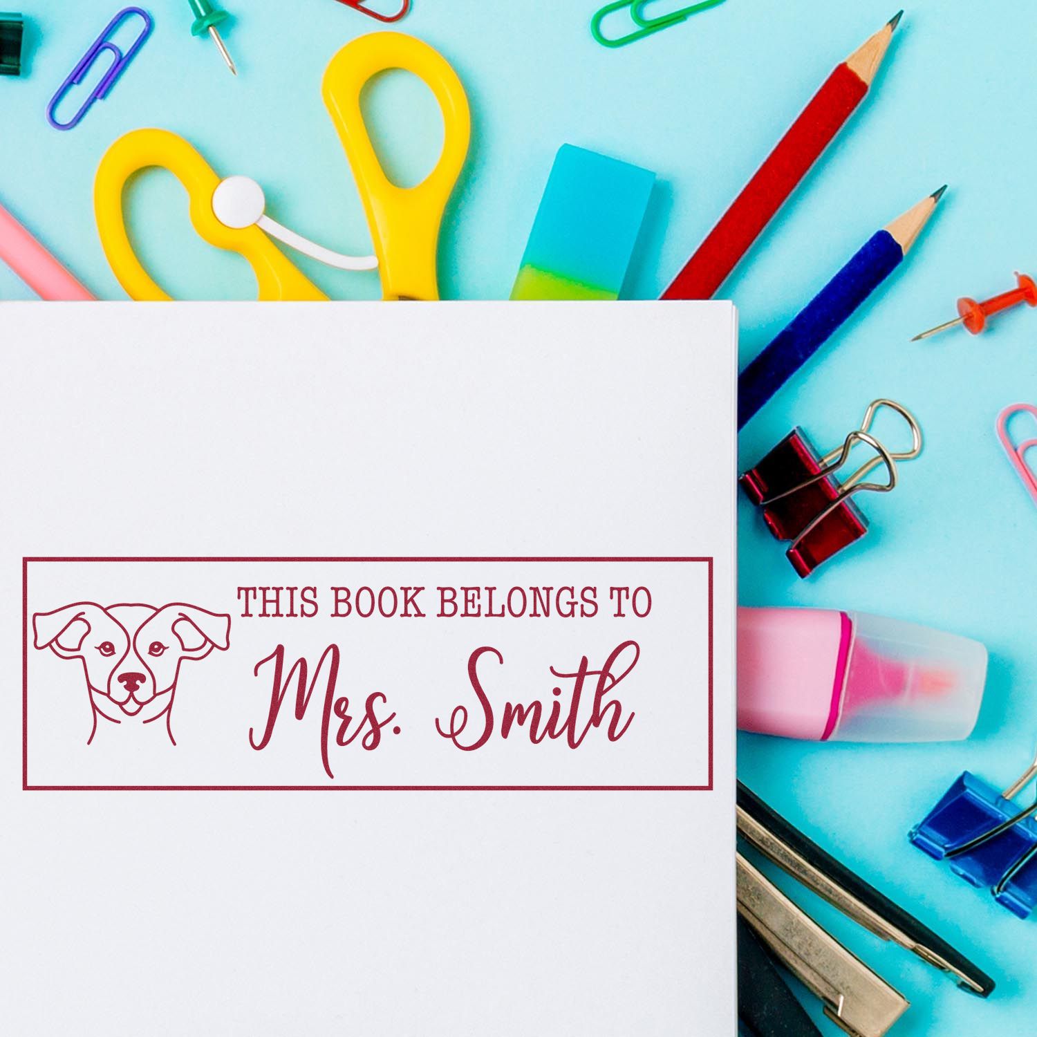 Custom rubber book stamp with dog icon and "This Book Belongs to Mrs. Smith" on white paper with school supplies.