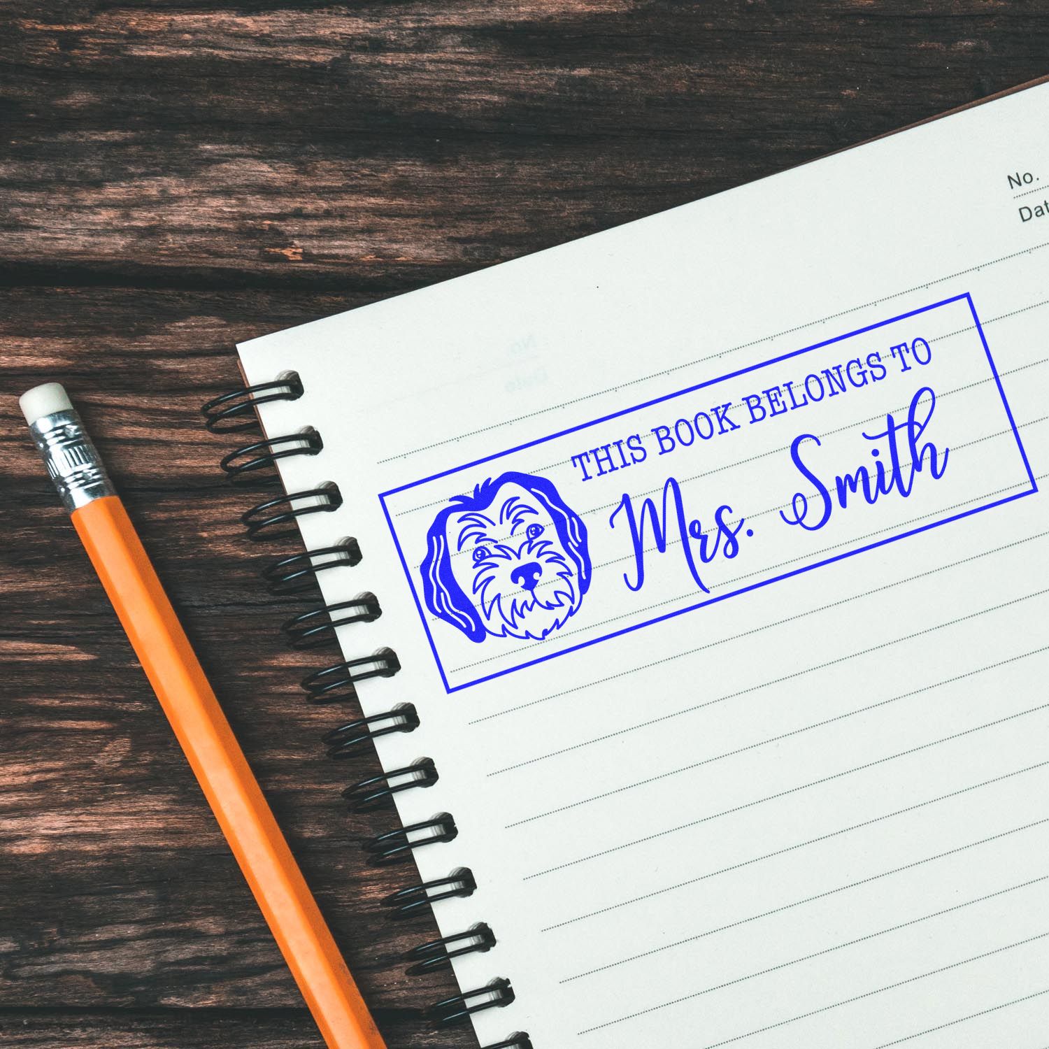 Personalized rubber book stamp with dog icon, marking This Book Belongs to Mrs. Smith on a notebook.