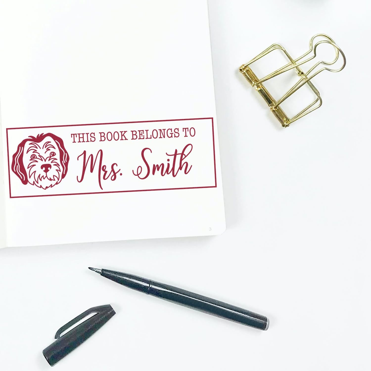 Personalized rubber book stamp for Mrs. Smith with dog design, marking a bookplate on a page with binder clip and pen.