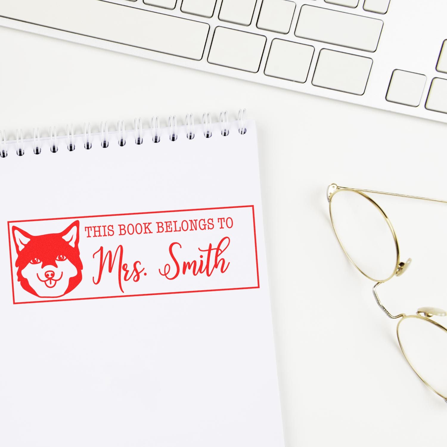 Personalized rubber book stamp reading This book belongs to Mrs. Smith with dog icon, shown on a notebook beside keyboard and glasses.