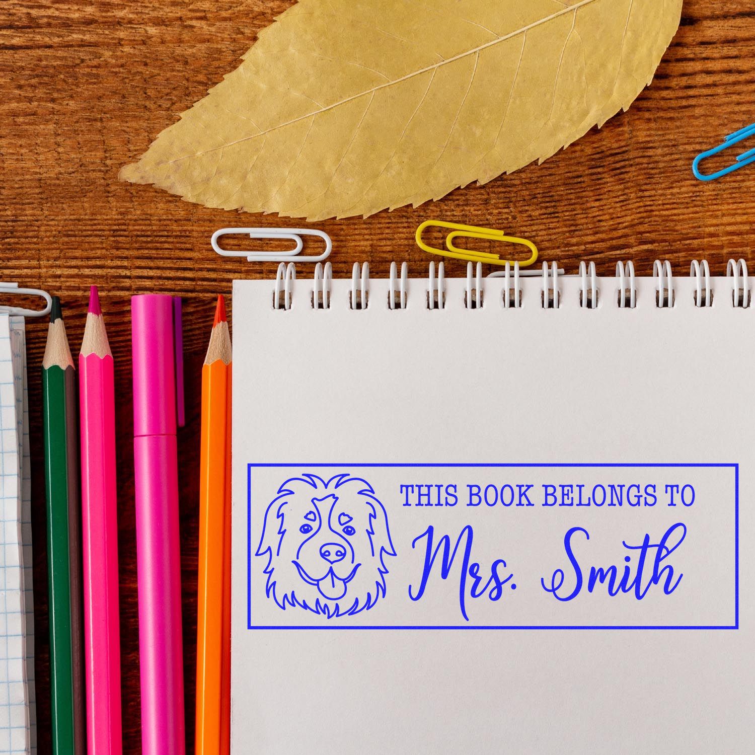 Personalized rubber book stamp with dog design reading This Book Belongs to Mrs. Smith, shown on a notebook with school supplies.