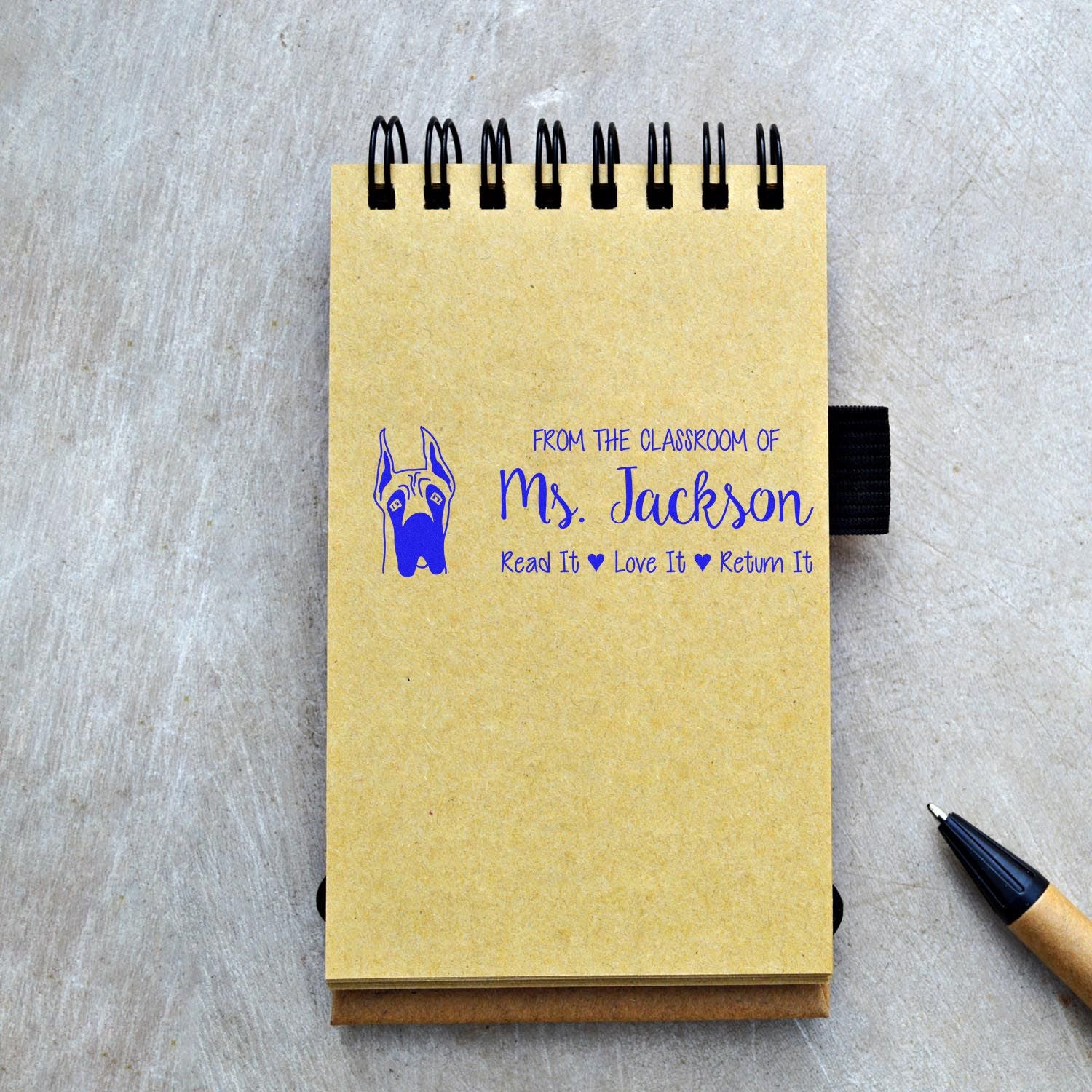 Personalized teacher rubber stamp on notebook: From the classroom of Ms. Jackson, Read It, Love It, Return It with dog icon.