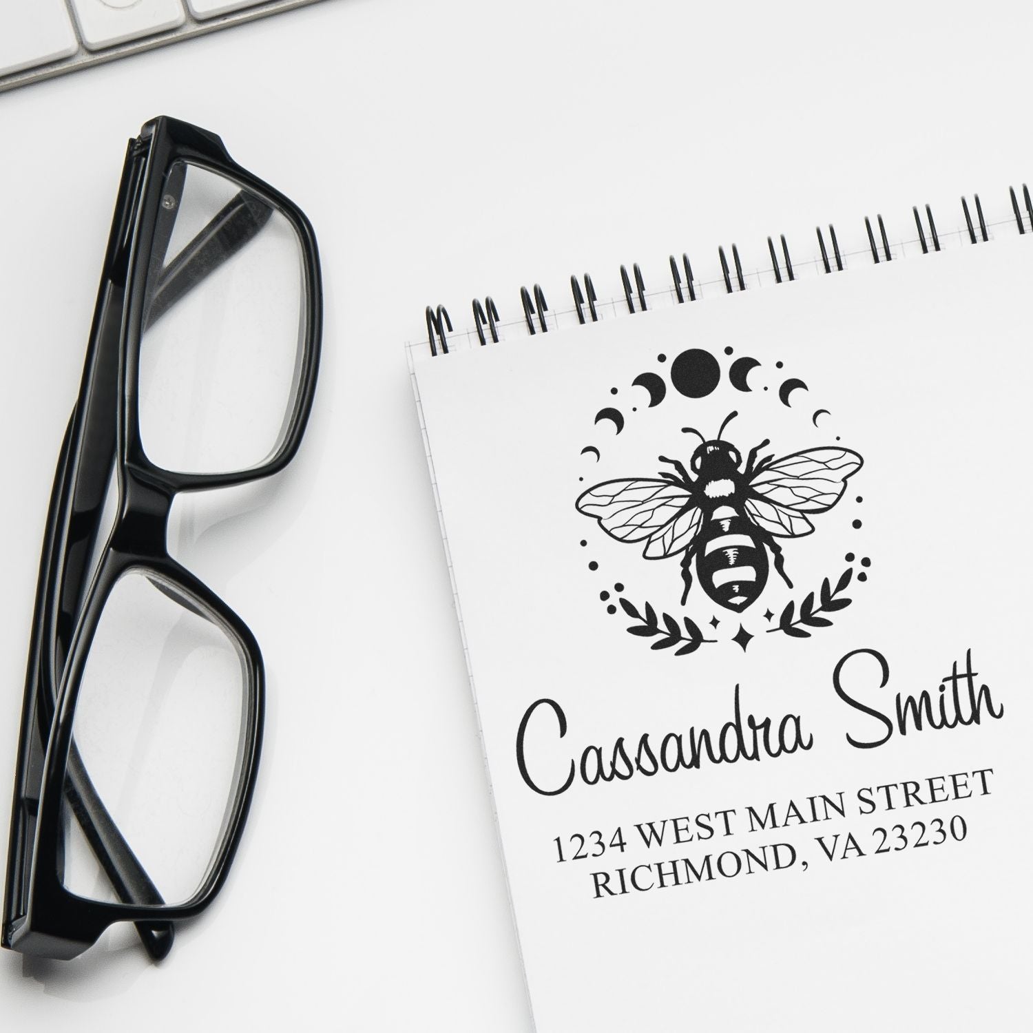 Personalized bee return address rubber stamp design with name and address on a notebook beside black eyeglasses.