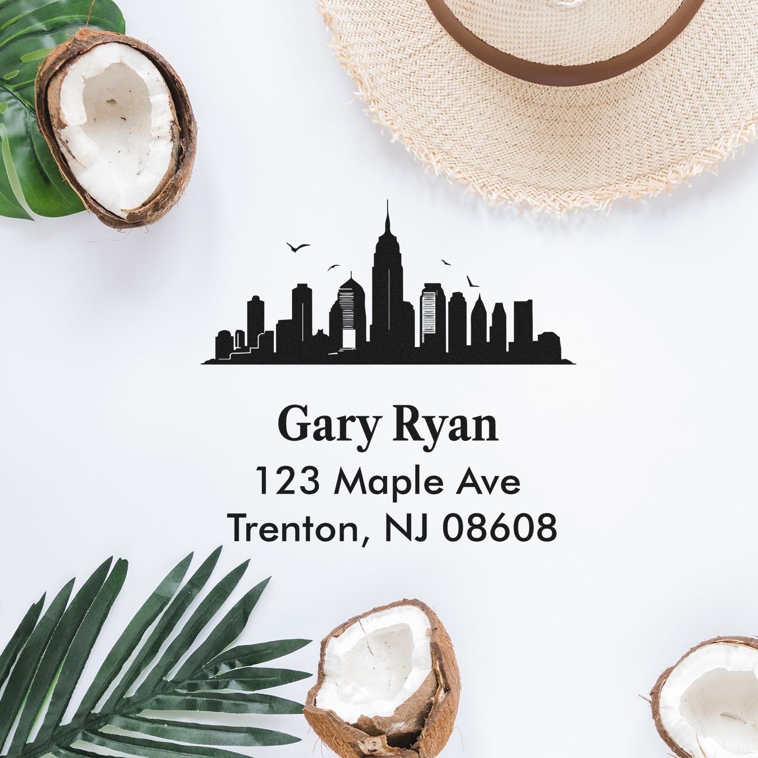 Custom address rubber stamp with skyline logo, Gary Ryan, 123 Maple Ave, Trenton NJ 08608, styled on tropical flat lay.