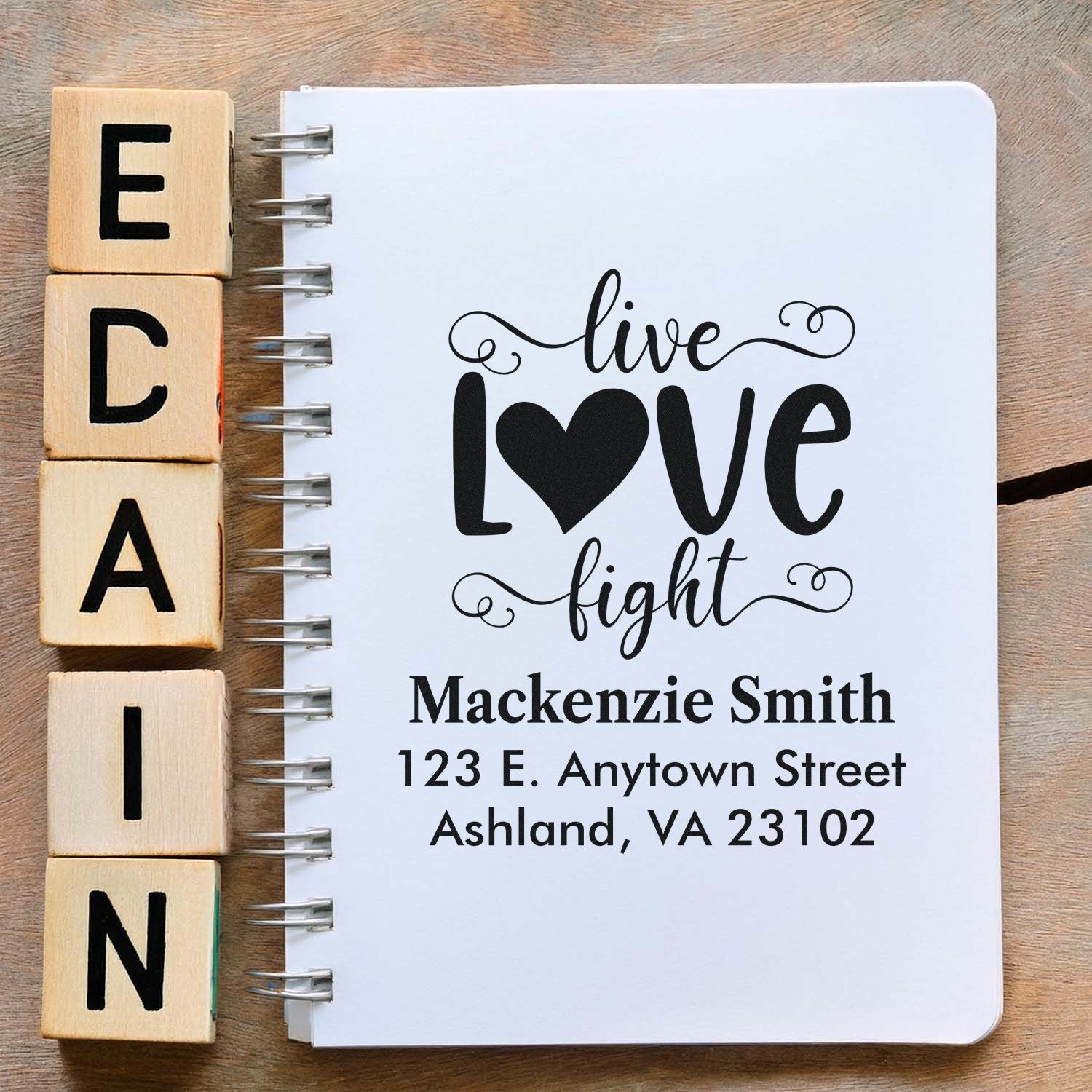 Wooden alphabet rubber stamps beside a notebook featuring a custom address stamp design with live love fight and personalized address.