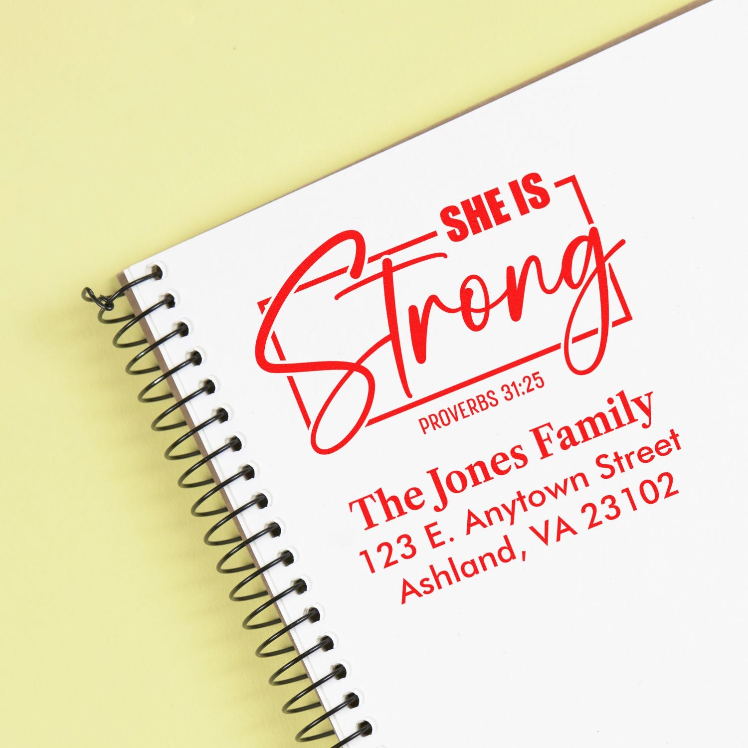 Custom rubber stamp impression reading She Is Strong Proverbs 31:25 with return address on a spiral notebook.