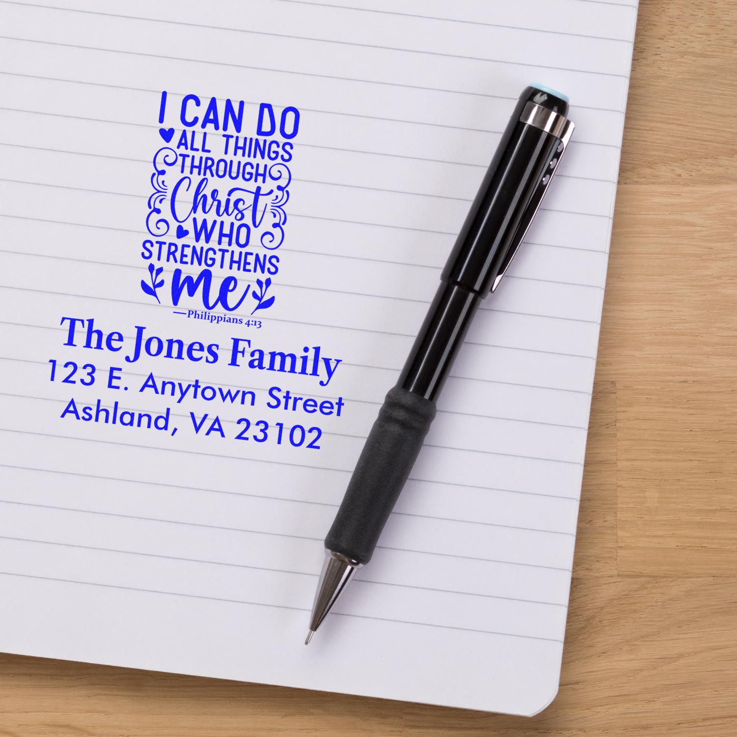 Custom Christian rubber stamp with Bible verse and family address stamped on a notebook beside a pen.