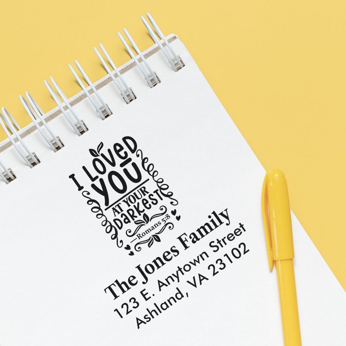 Custom rubber stamp examples: Christian quote and personalized family address stamp on a notebook with a yellow pen.