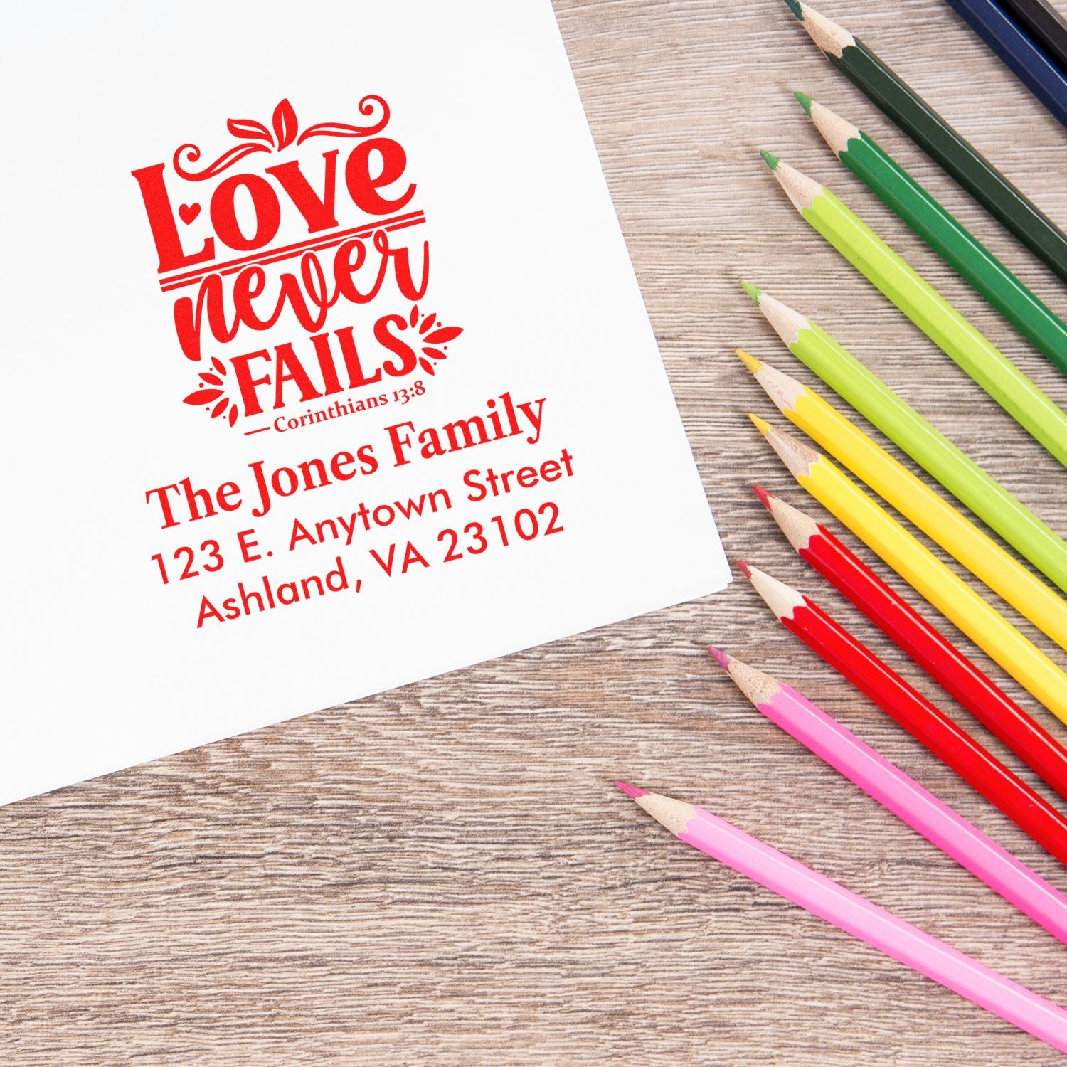 Custom rubber stamp with Love never fails and family return address printed on paper beside colorful pencils.