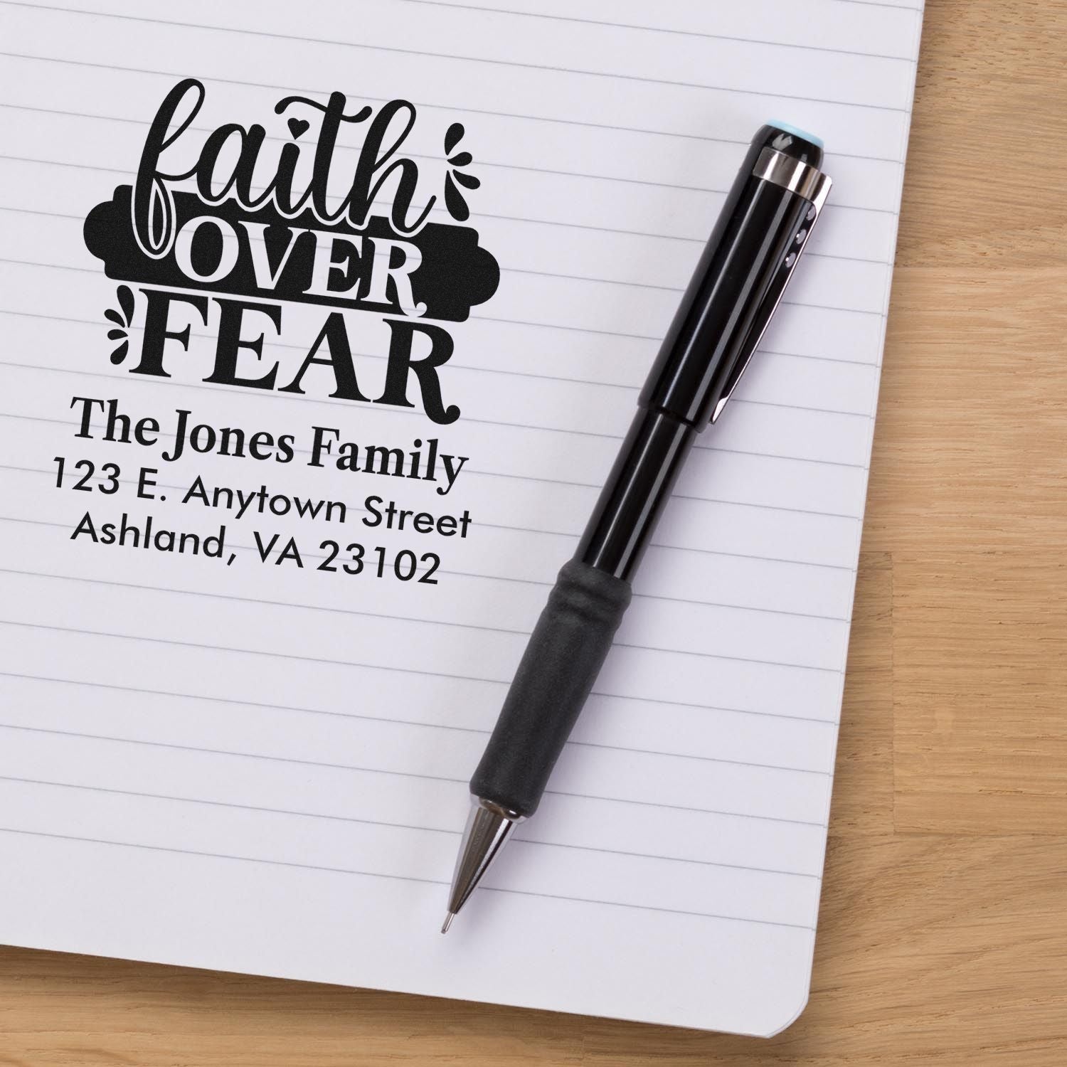 Custom rubber stamp imprint with Faith Over Fear and family return address on a notebook, beside a pen.