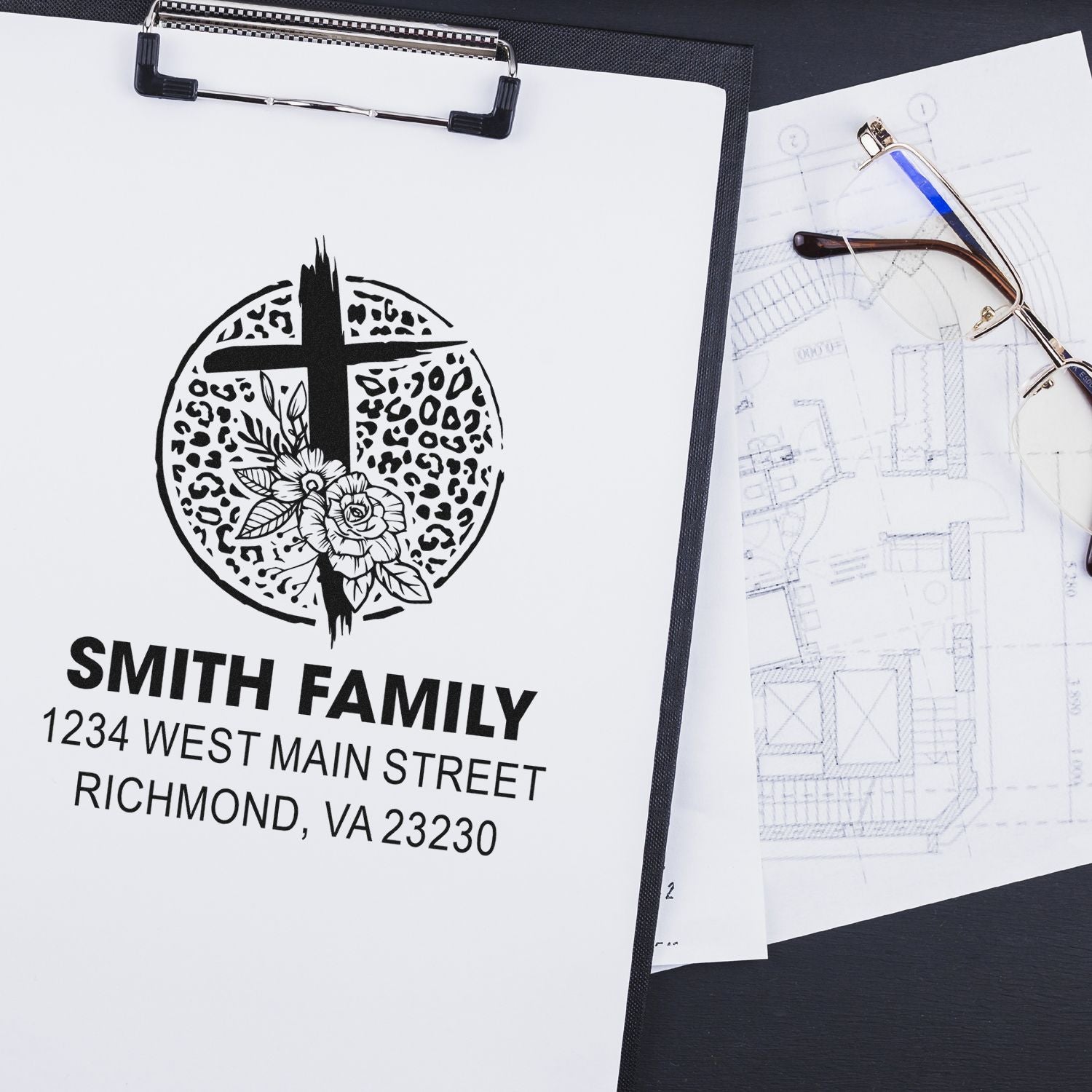 Custom family address rubber stamp with Smith Family floral cross design on paper beside blueprints and eyeglasses.